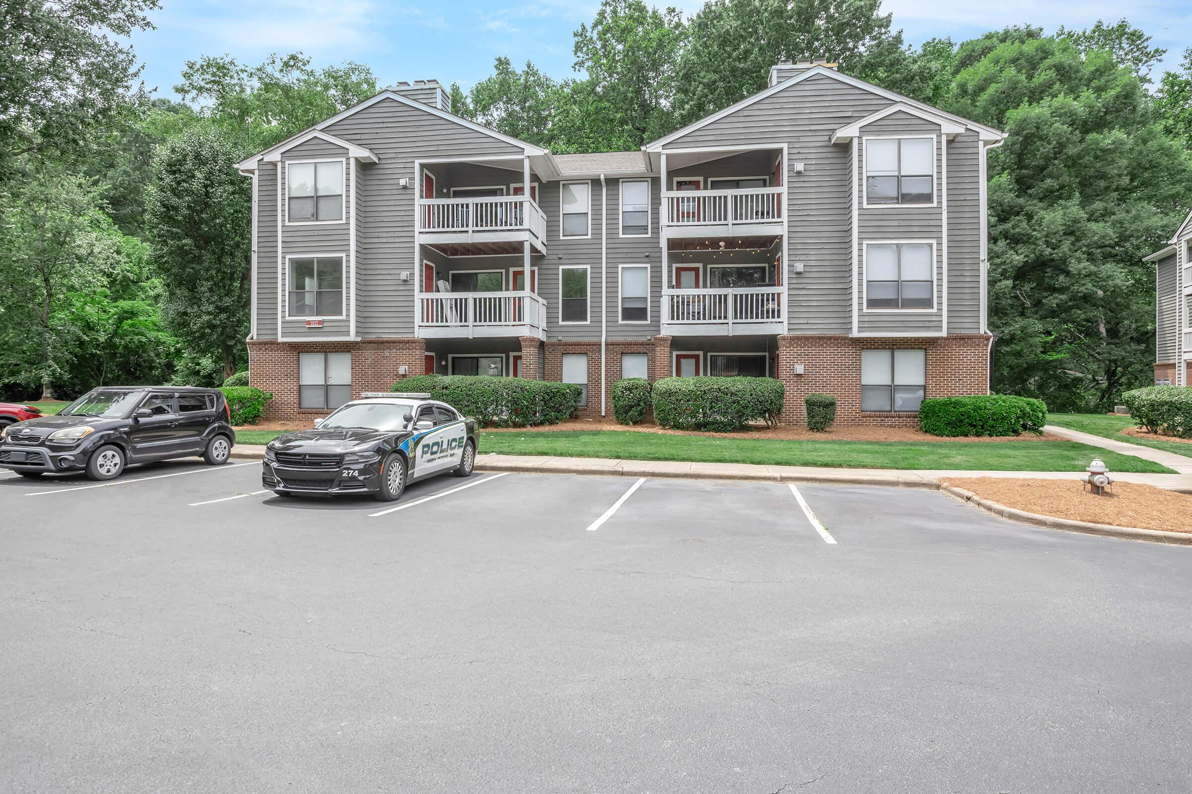 A three-story apartment building with gray siding and balconies, surrounded by trees and landscaped areas. In the foreground, a police car is parked alongside several empty parking spaces, and two other vehicles are visible in the lot. The scene is set on a sunny day.