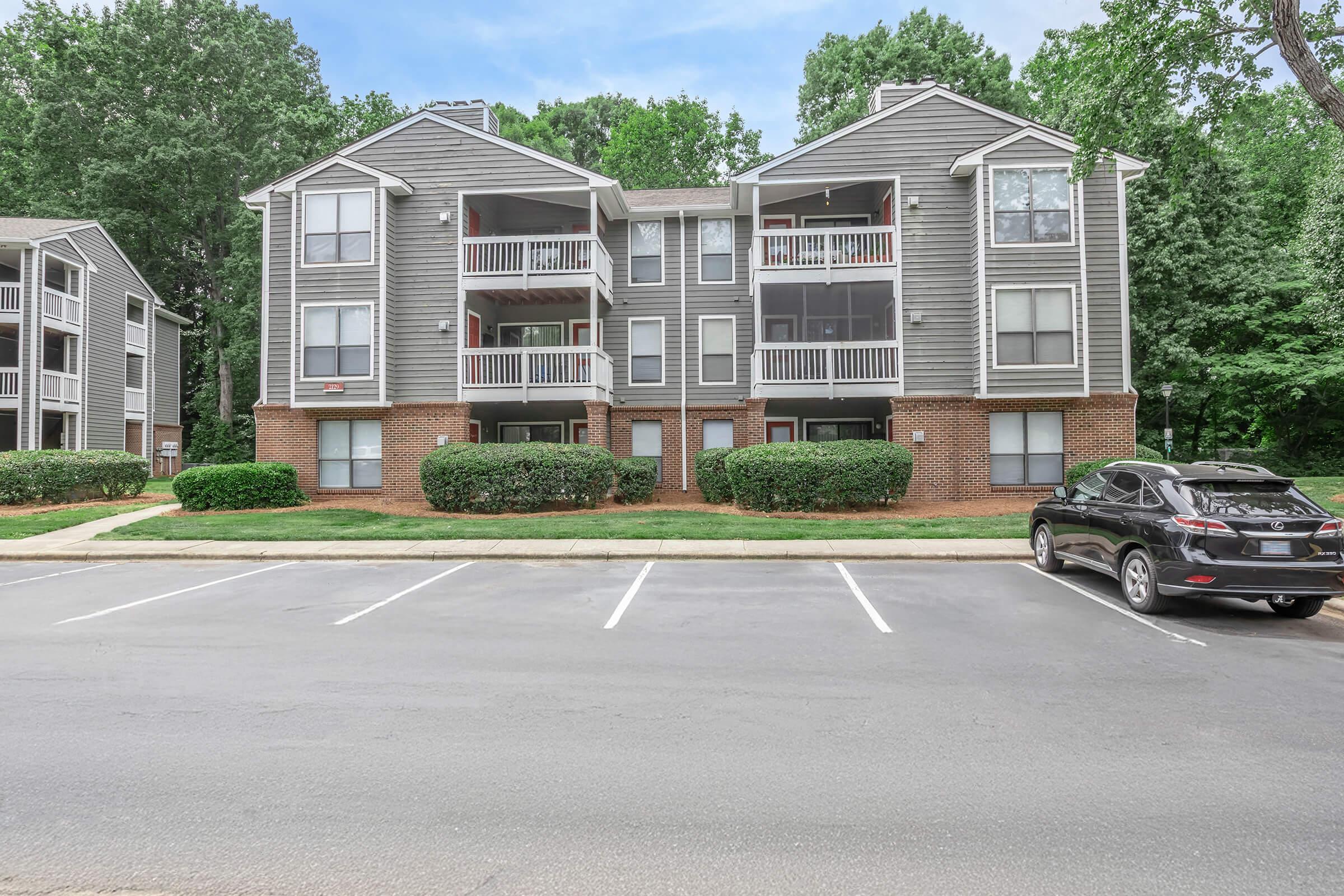 Multifamily apartment building with three floors, featuring balconies on each unit. The building is surrounded by lush greenery and shrubs, and there is a parking lot in the foreground with several open spaces.