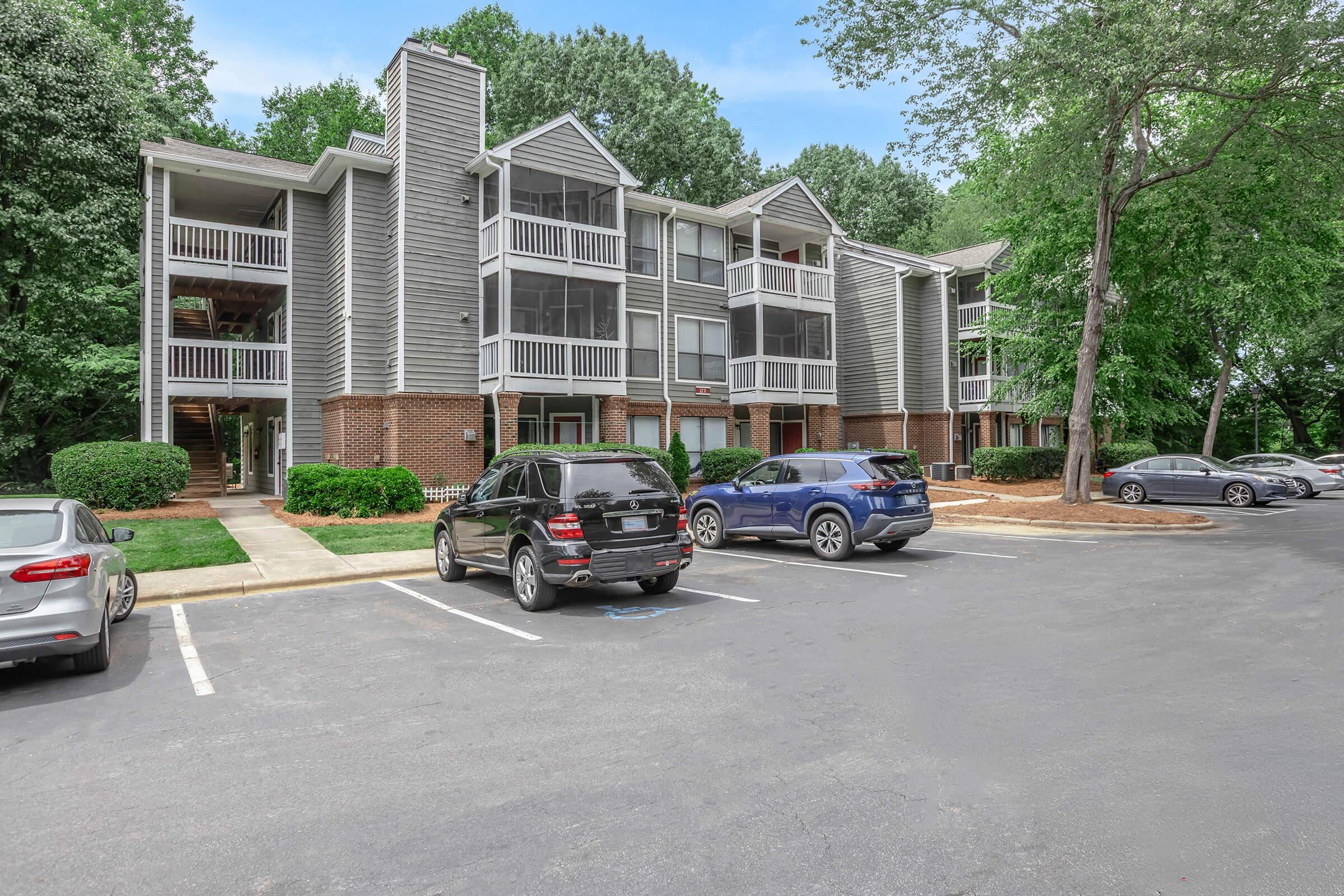 A residential apartment building with three stories, featuring multiple balconies and a mix of brick and siding exterior. The surroundings include green trees and well-maintained shrubbery. Several parked cars are visible in the foreground on a paved parking lot.