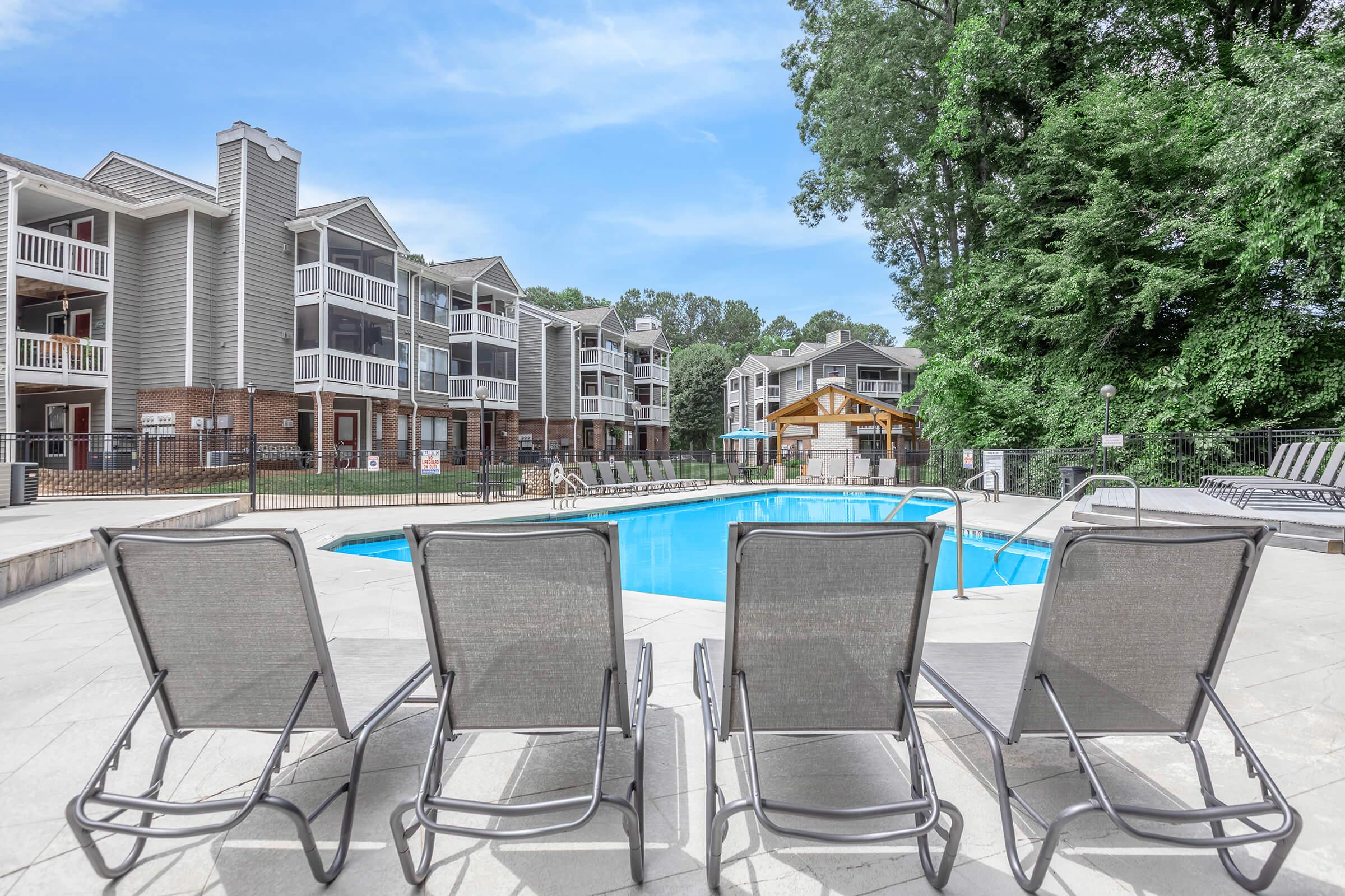 A serene outdoor pool area featuring several lounge chairs in the foreground, surrounded by well-maintained landscaping and residential buildings in the background. The sky is clear, and the pool is inviting, creating a relaxing atmosphere for residents.