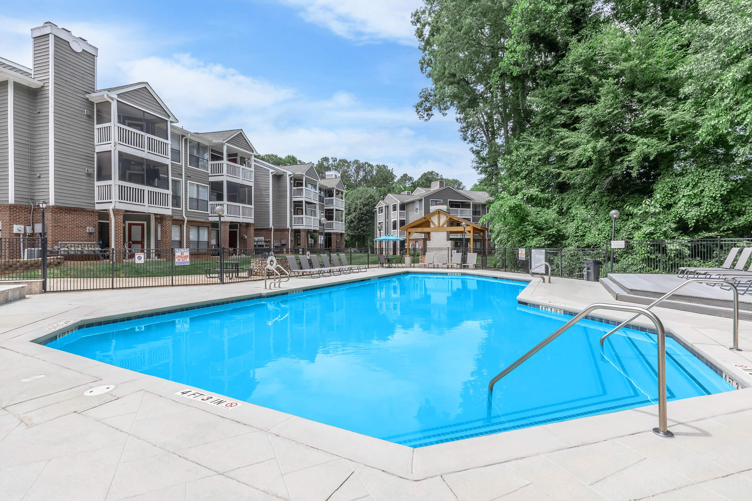 A clear blue swimming pool surrounded by lounge chairs and a fence, with well-maintained apartment buildings in the background. Lush green trees provide shade, and a shaded gazebo is visible nearby, creating a relaxing outdoor atmosphere.