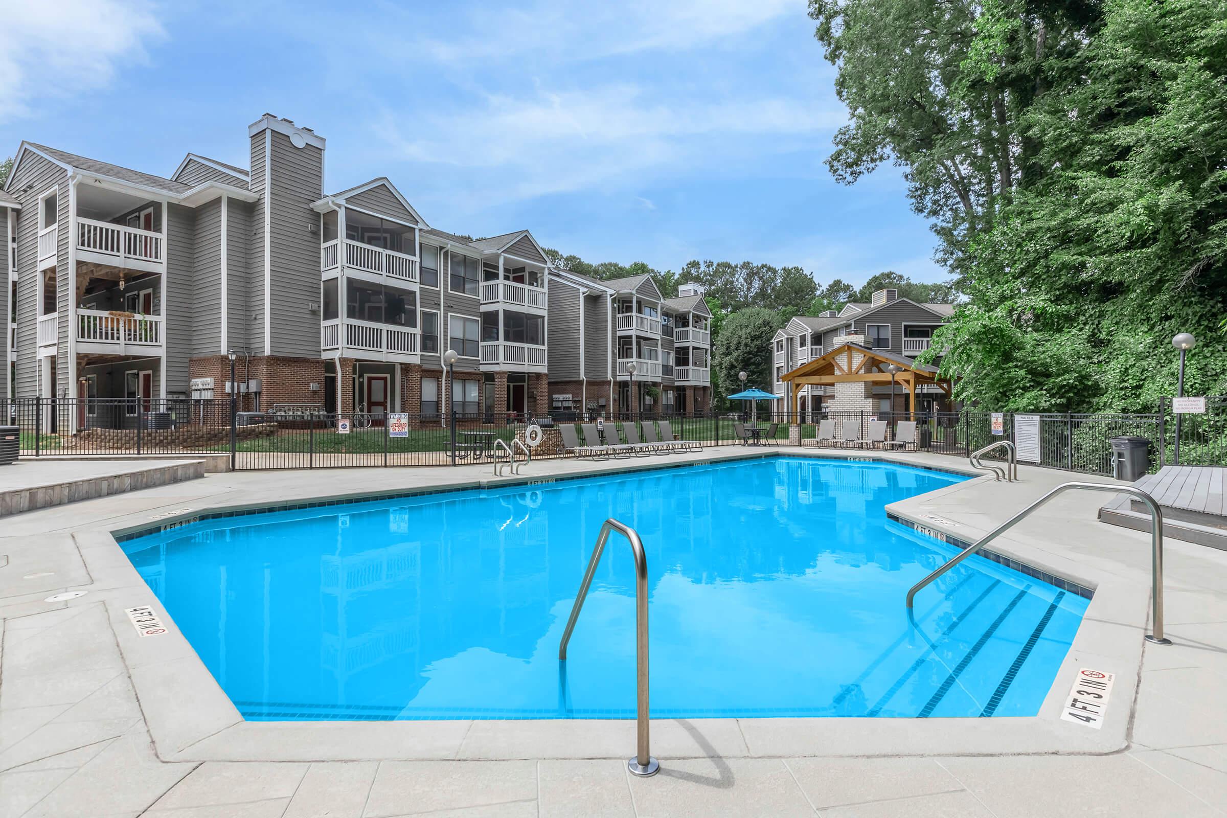 A vibrant outdoor swimming pool surrounded by well-maintained apartments. The pool features a set of stairs for entry, with lounge chairs and a gazebo nearby. Lush greenery fills the background, creating a serene atmosphere ideal for relaxation and leisure.