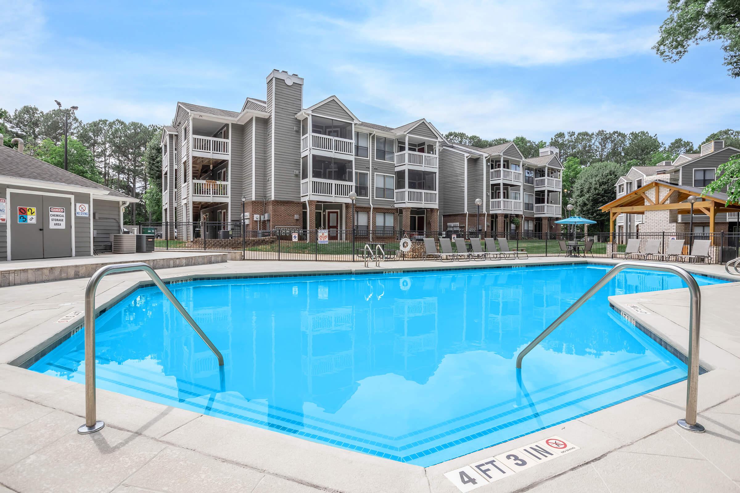A clear blue swimming pool with steps and handrails, surrounded by a grassy area and buildings in the background. The buildings have multiple levels with balconies, and there is a shaded seating area nearby. Trees and a bright sky enhance the inviting atmosphere.