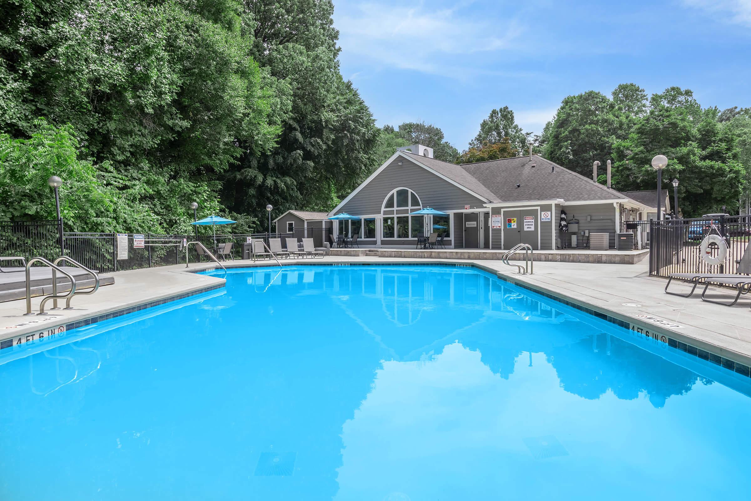 A clear blue swimming pool with lounge chairs around it, surrounded by green trees. In the background, a modern building serves as a pool house, featuring large windows and outdoor seating areas. The pool area is well-maintained and inviting under a bright blue sky.