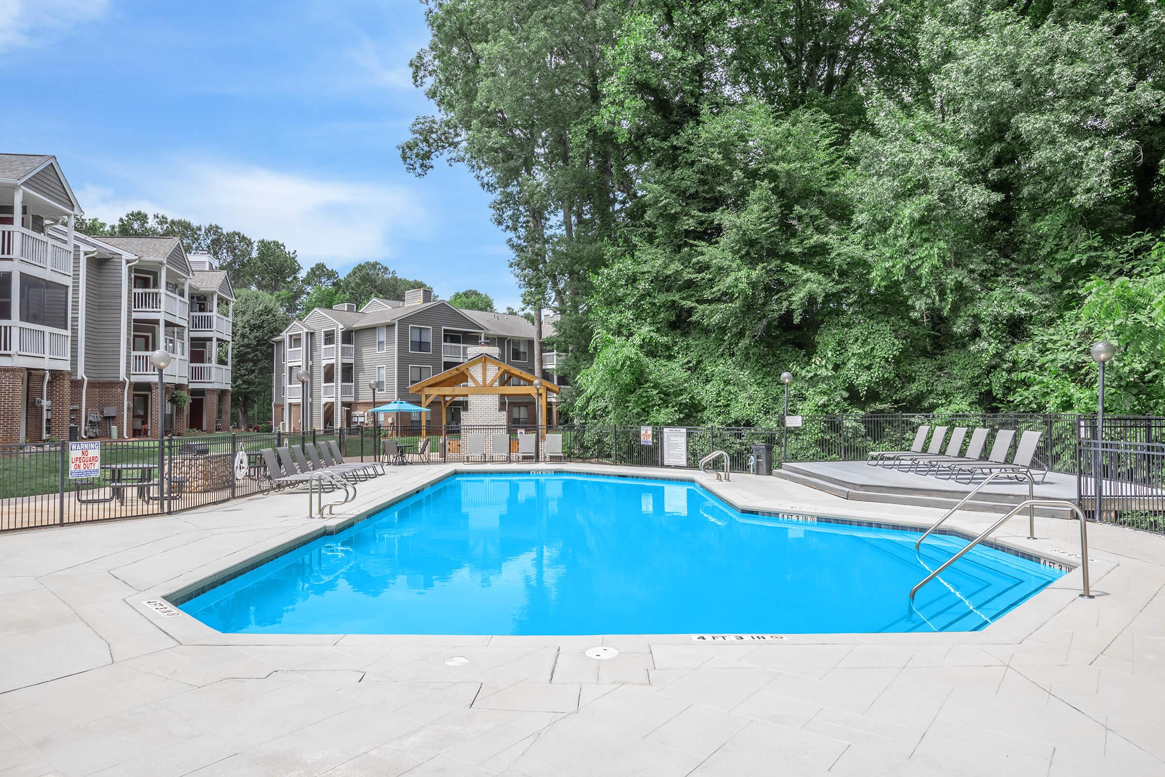 A clean, blue swimming pool surrounded by a patio area with lounge chairs. In the background, several apartment buildings are nestled among lush greenery. A gazebo with picnic tables is also visible near the pool, creating a welcoming outdoor space for relaxation and leisure.