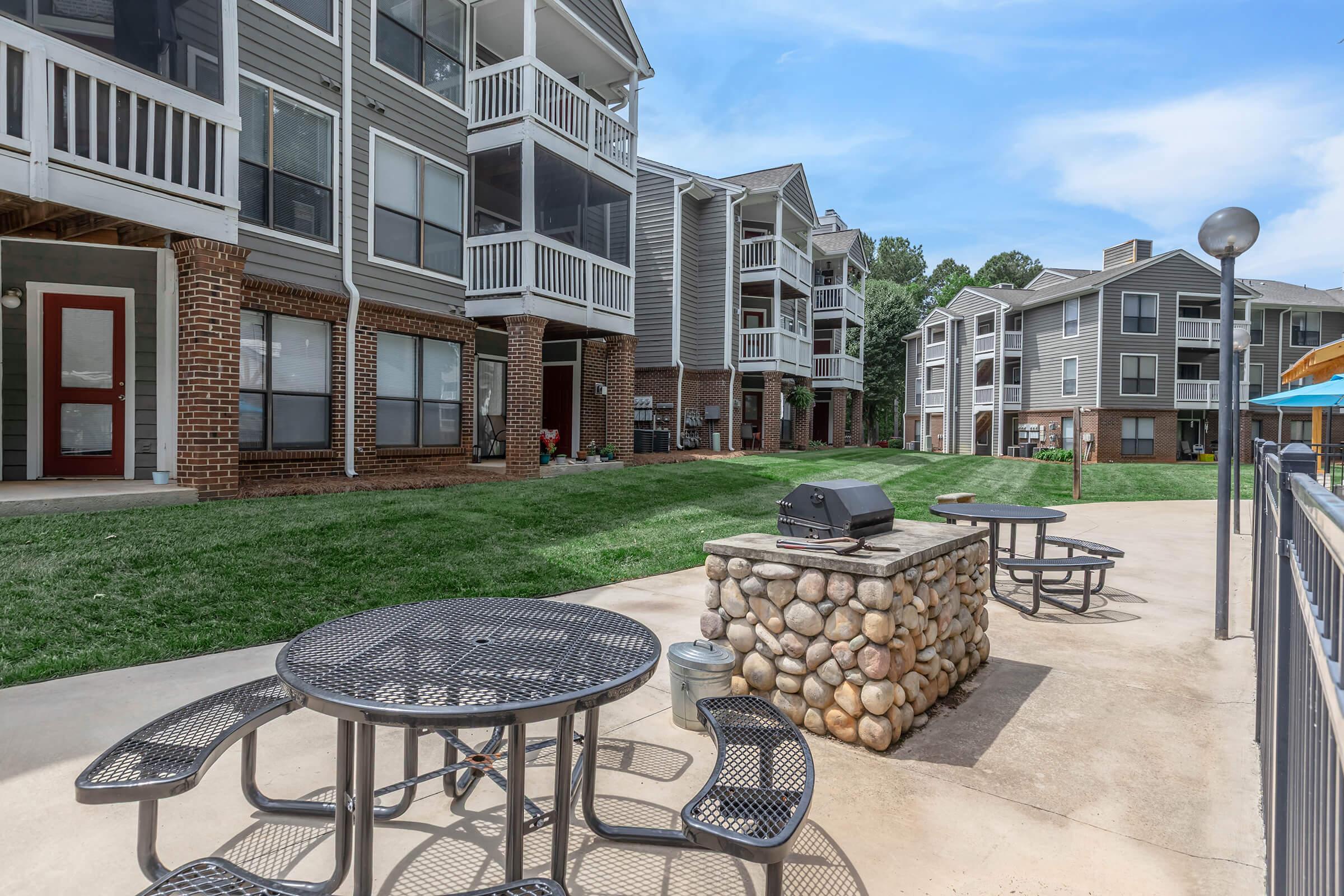 A view of a landscaped courtyard featuring a barbecue grill on a stone pedestal, surrounded by picnic tables. In the background, several multi-story apartment buildings with balconies and a well-maintained lawn are visible under a clear blue sky.