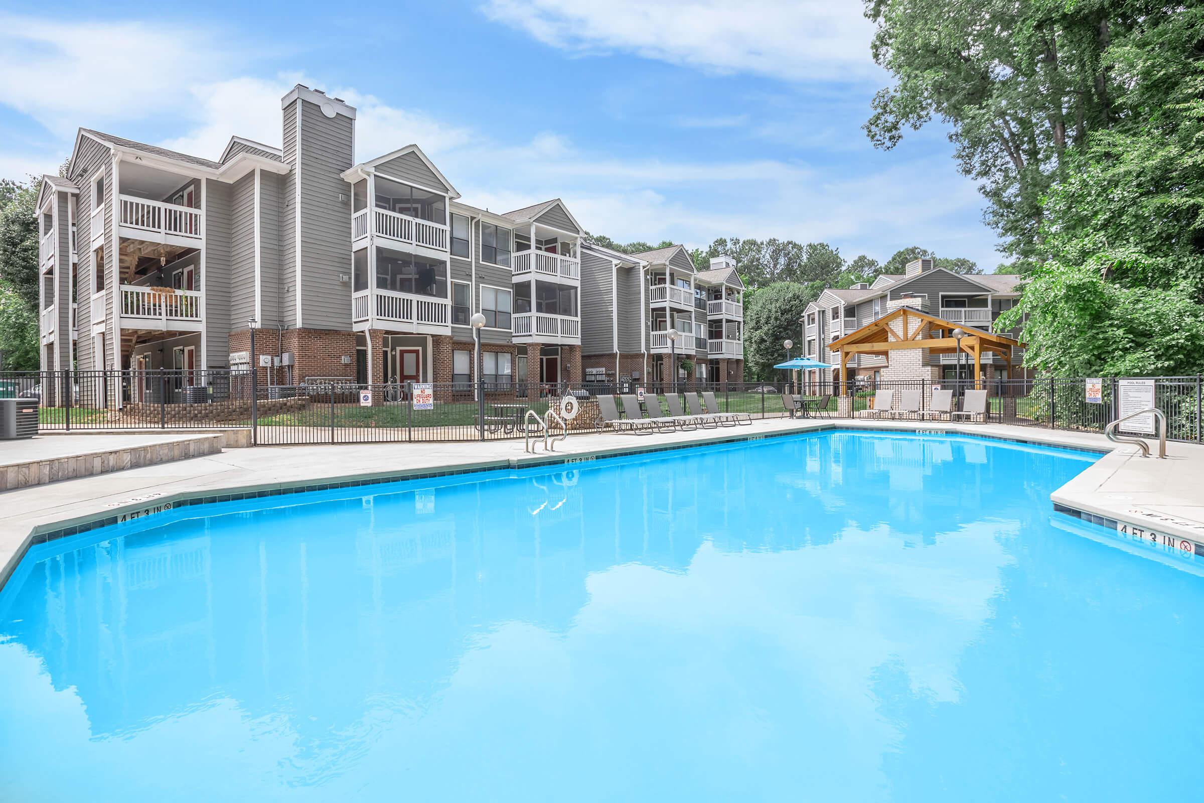 A clear blue swimming pool in the foreground, bordered by a spacious patio. In the background, there are several multi-story apartment buildings with balconies and greenery surrounding the area. A covered gazebo is visible near the pool, adding to the outdoor amenities. Trees provide shade in the vicinity.