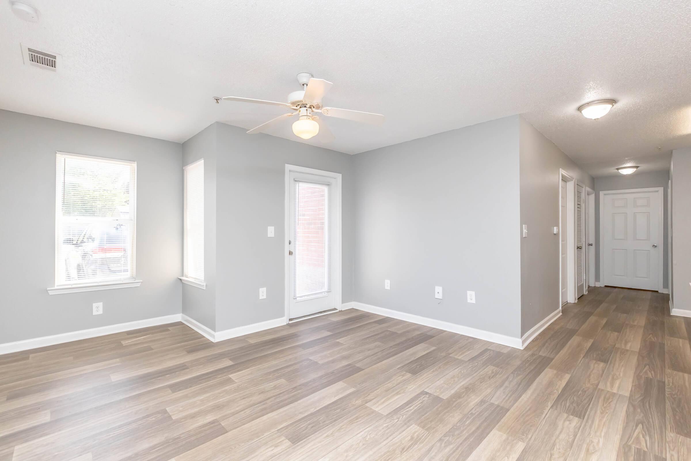 A spacious interior of an apartment featuring light gray walls, wood-like flooring, and a ceiling fan. There are two windows allowing natural light, a door leading outside, and a hallway visible at the far end with closed doors on either side. The room appears bright and inviting.
