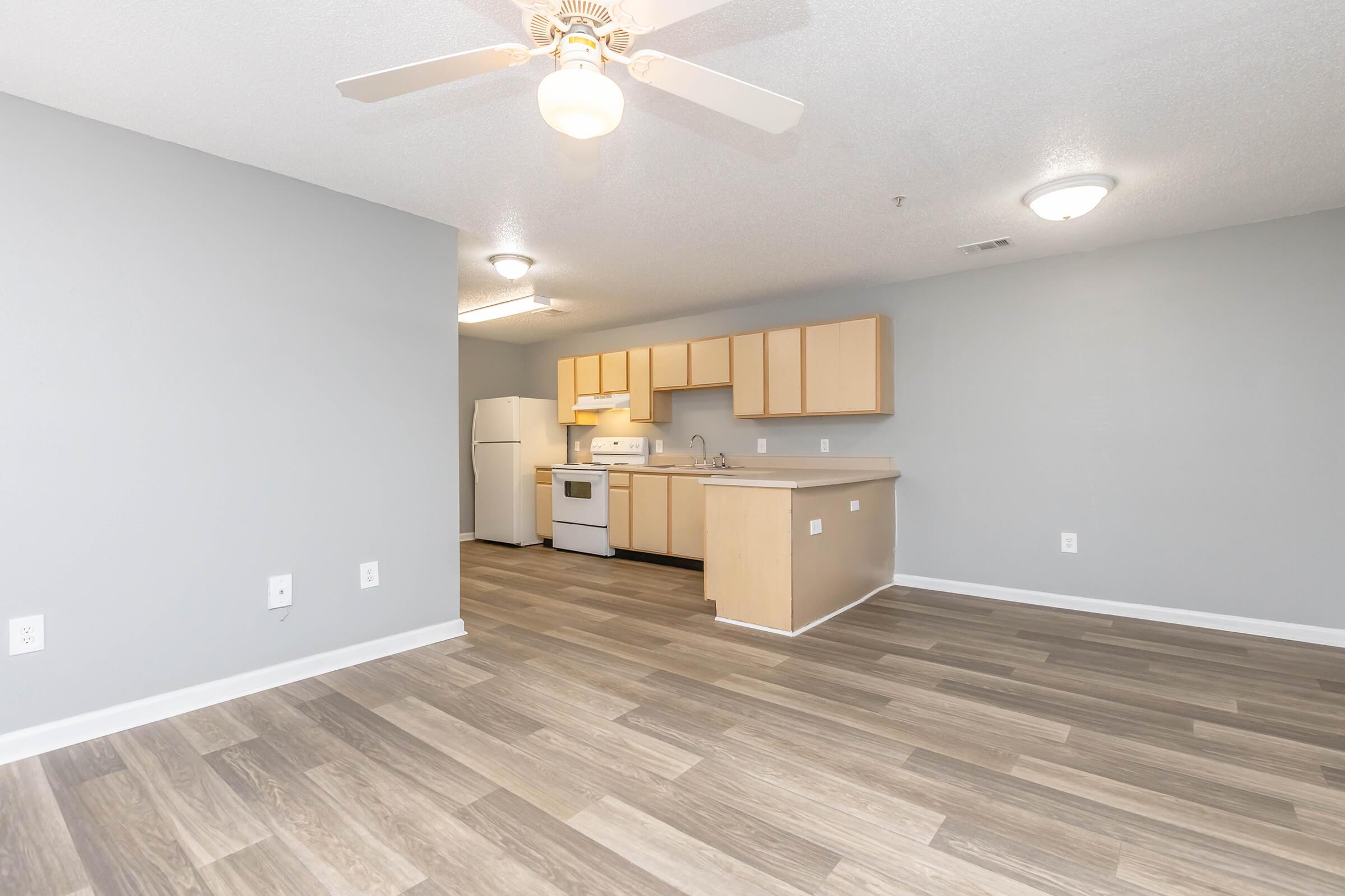 Spacious kitchen and living area in a modern apartment. Light gray walls complement the wood-style flooring. The kitchen features wooden cabinets, a white refrigerator, and an oven. A ceiling fan hangs above, and natural light illuminates the space from overhead fixtures.