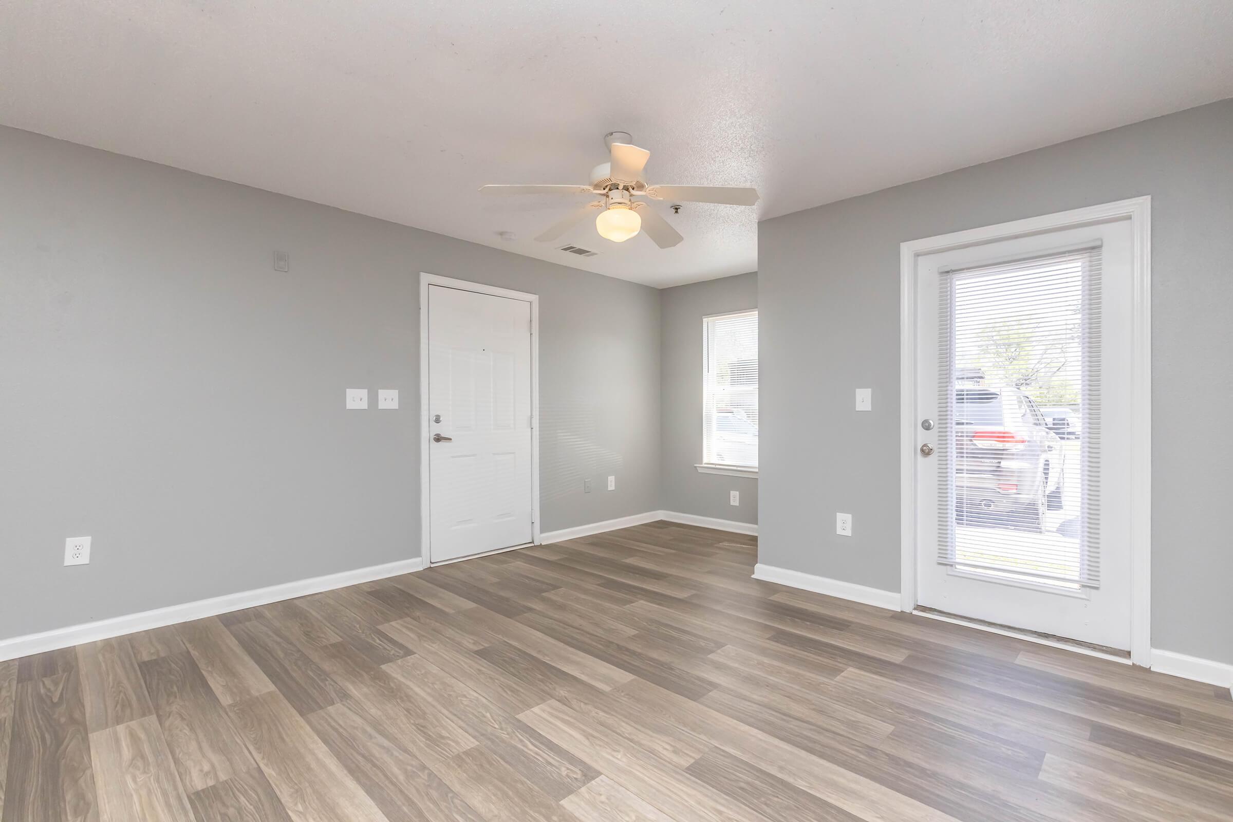 A bright, empty living room featuring gray walls and light brown wood-style flooring. There is a ceiling fan, a door leading outside, and a second door that appears to be a front entry. Natural light enters from a window on the right.