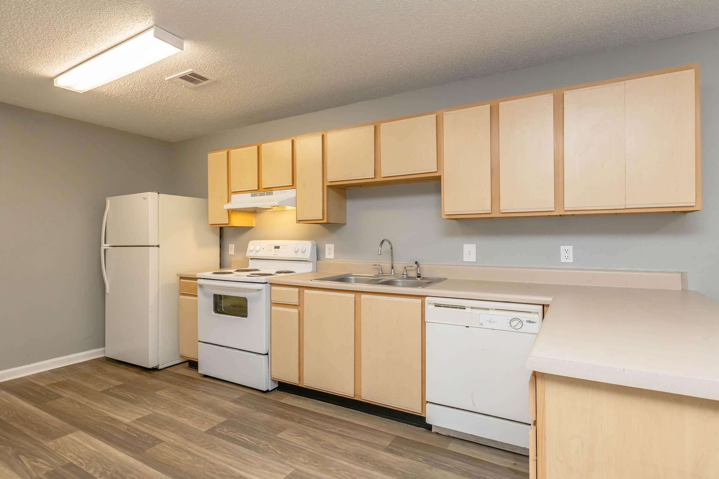 A modern kitchen featuring light wood cabinets, a white refrigerator, an oven, and a dishwasher. The countertop is light-colored and there is a sink with a faucet. The walls are painted gray, and the flooring is made of dark wood. Ceiling lights provide adequate lighting in the space.