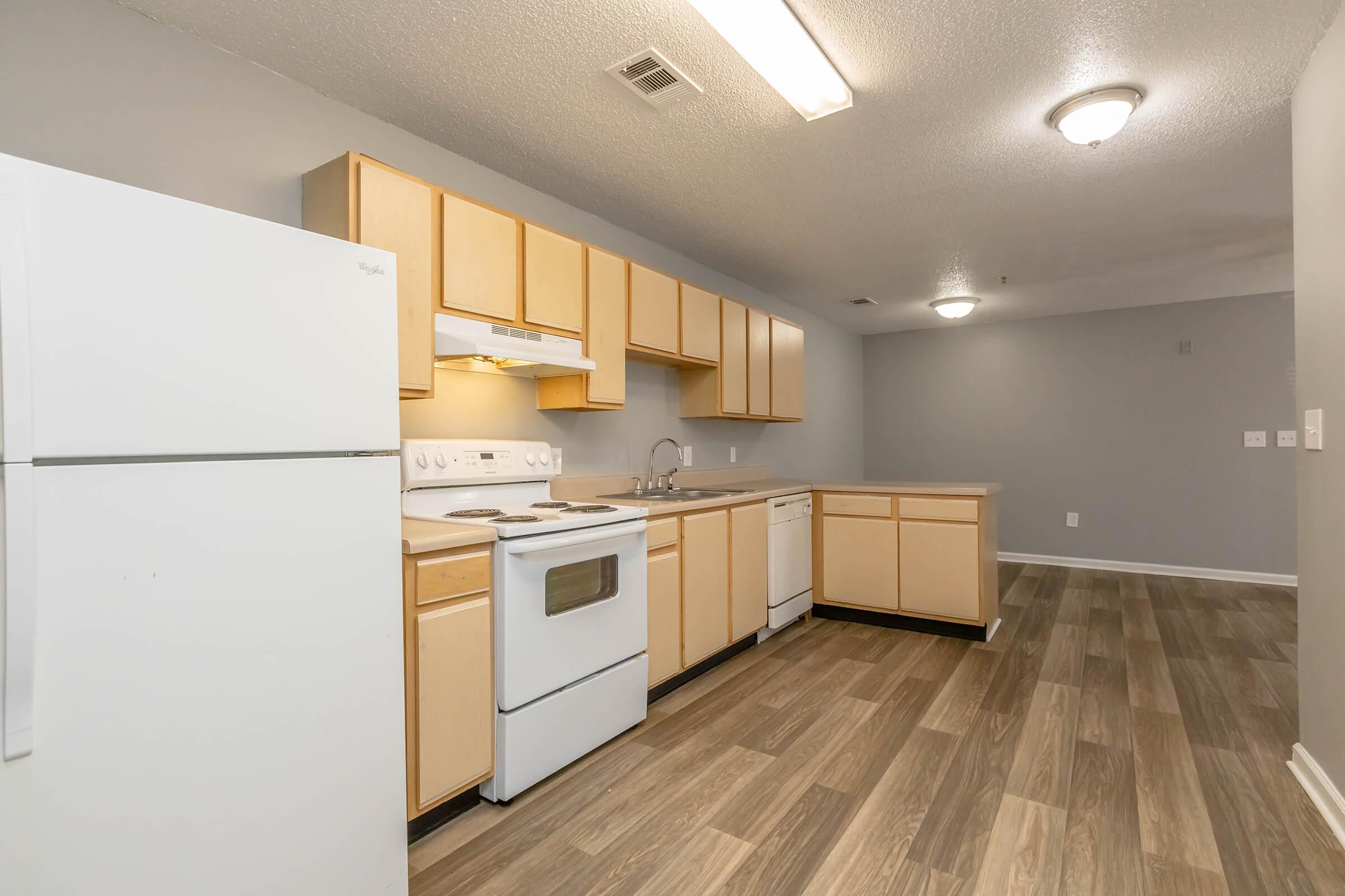 A modern kitchen featuring light wood cabinetry, a white refrigerator, stove, and dishwasher, with a spacious layout. The floor is covered in wood-like vinyl, and there is overhead lighting, creating a bright and inviting atmosphere.