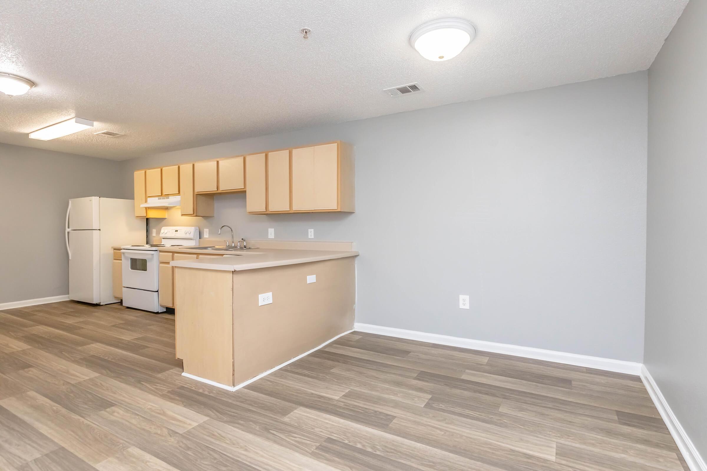 Modern kitchen and living area featuring light wood cabinetry, white appliances, and a neutral gray wall color. The space showcases laminate flooring and an open layout with ample natural light, creating a warm and inviting atmosphere.