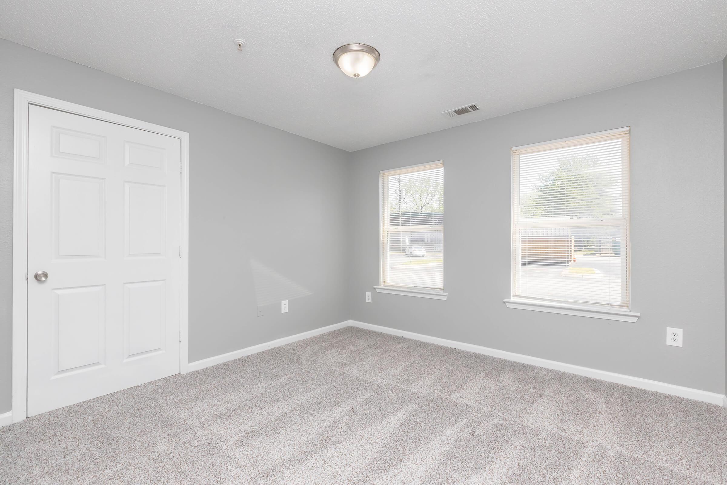 A well-lit empty bedroom featuring light gray walls, two windows with blinds, and soft carpeting. The room includes a white closet door and overhead light fixture, creating a clean and inviting space.