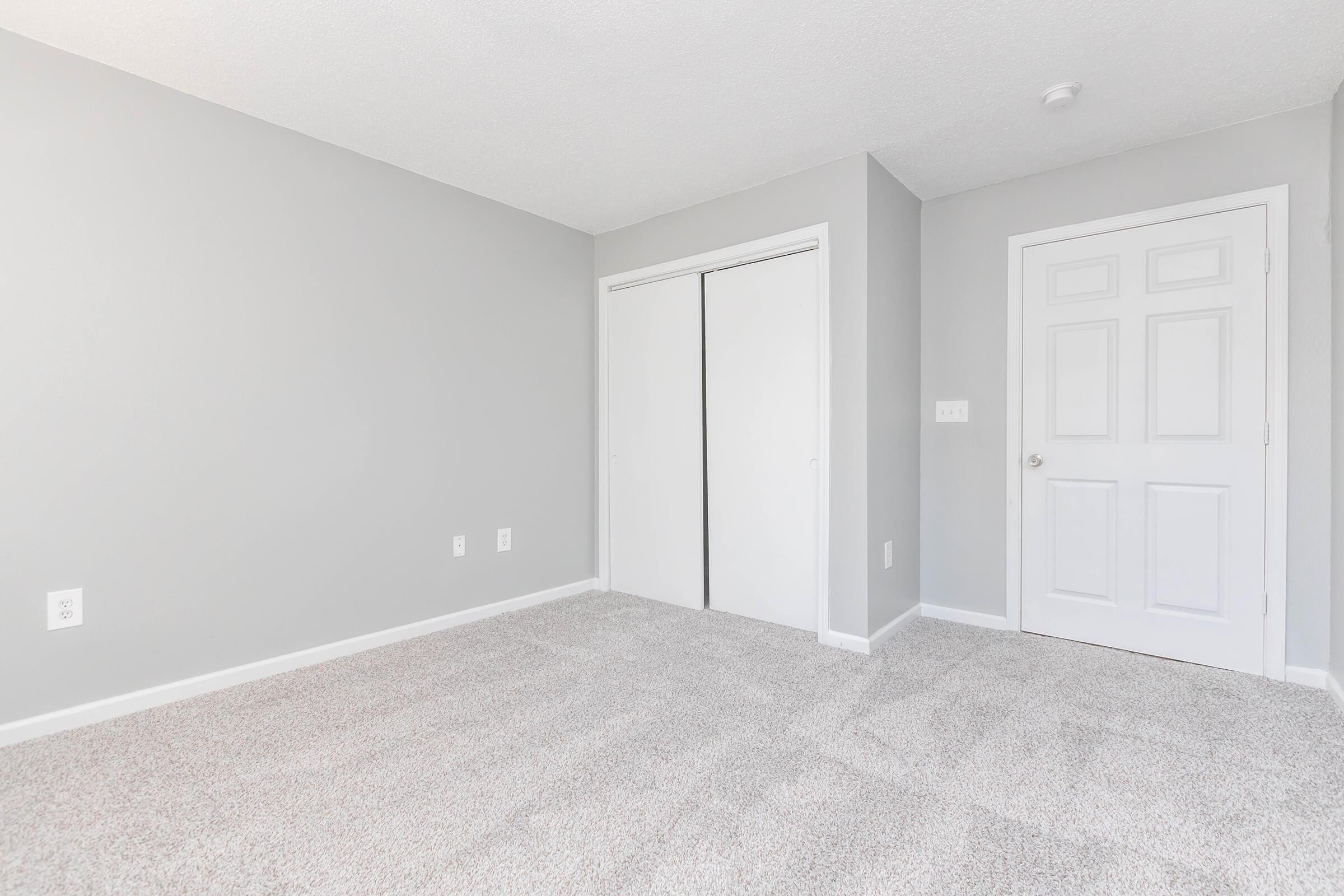 A spacious, empty bedroom featuring light gray walls, soft carpet flooring, and a closet with sliding doors. The room has a single door leading to another area, with natural light illuminating the space, creating a clean and fresh atmosphere.