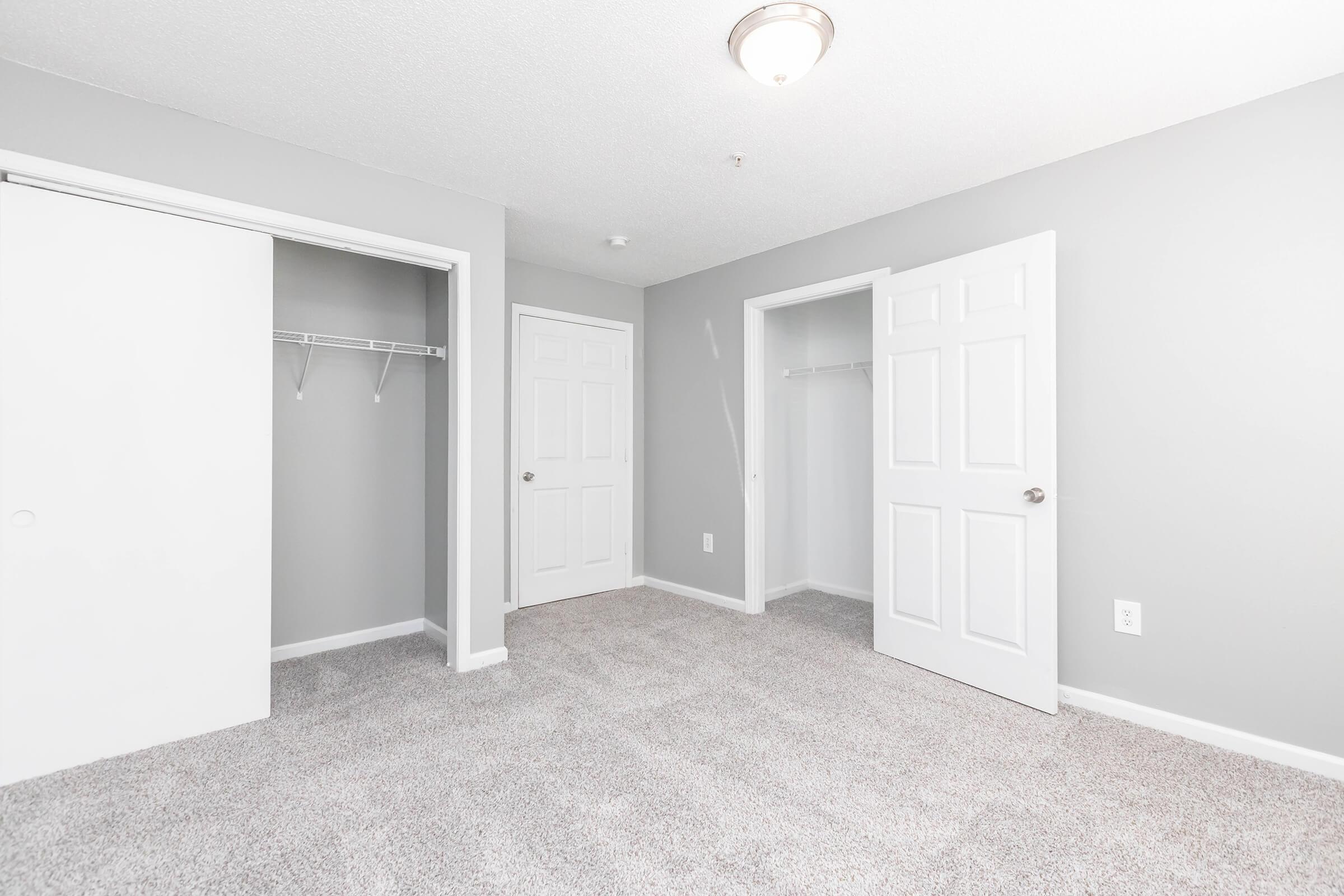 A spacious room with light gray walls, featuring two closets with sliding and hinged doors. The floor is covered in soft, light-colored carpet, and there’s a ceiling light fixture. The room appears well-lit and empty, suitable for various uses.