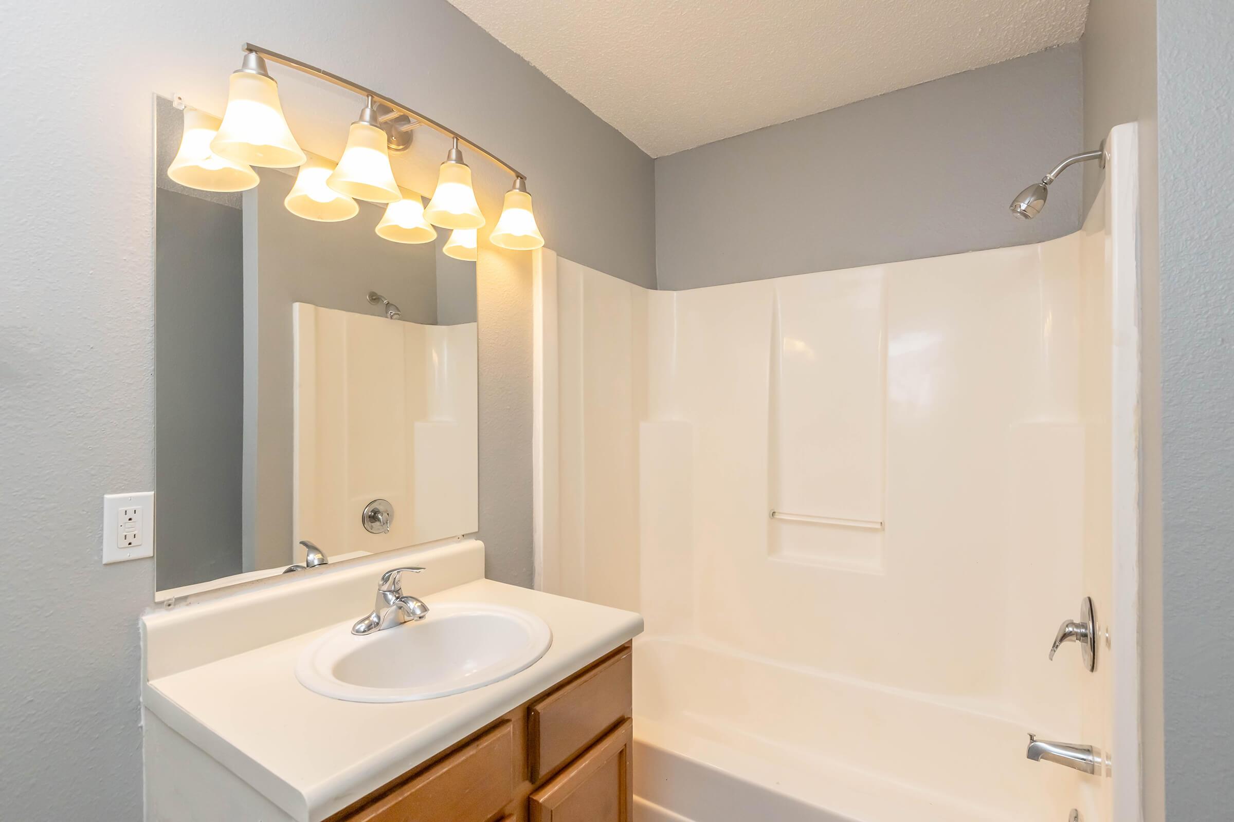 A clean bathroom featuring a white bathtub with a shower, a light-colored countertop with a sink, and a large mirror above it. Four pendant lights hang above the mirror, and the walls are painted a light gray, creating a modern and bright atmosphere.