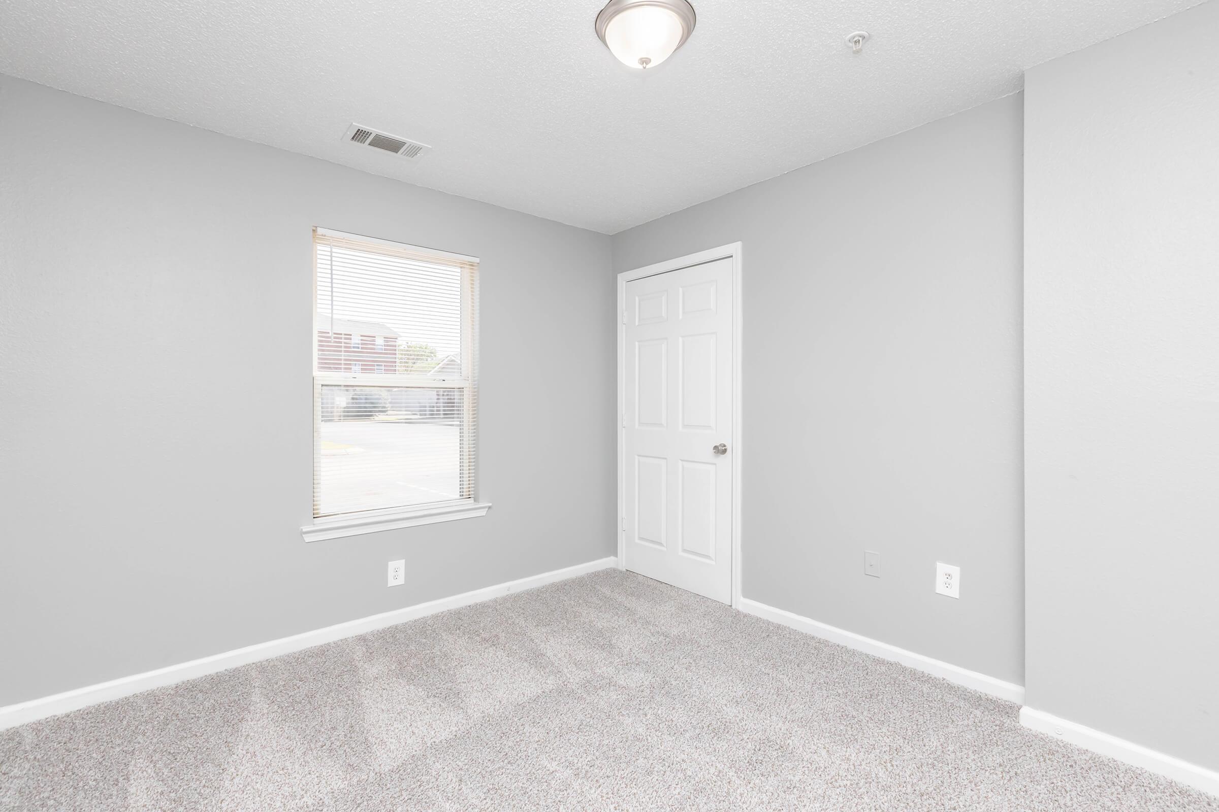 A small, vacant room with light gray walls and a carpeted floor. There is a window with blinds allowing natural light in, and a closed white door. The overall atmosphere is bright and minimalistic, suitable for various uses such as an office or bedroom.