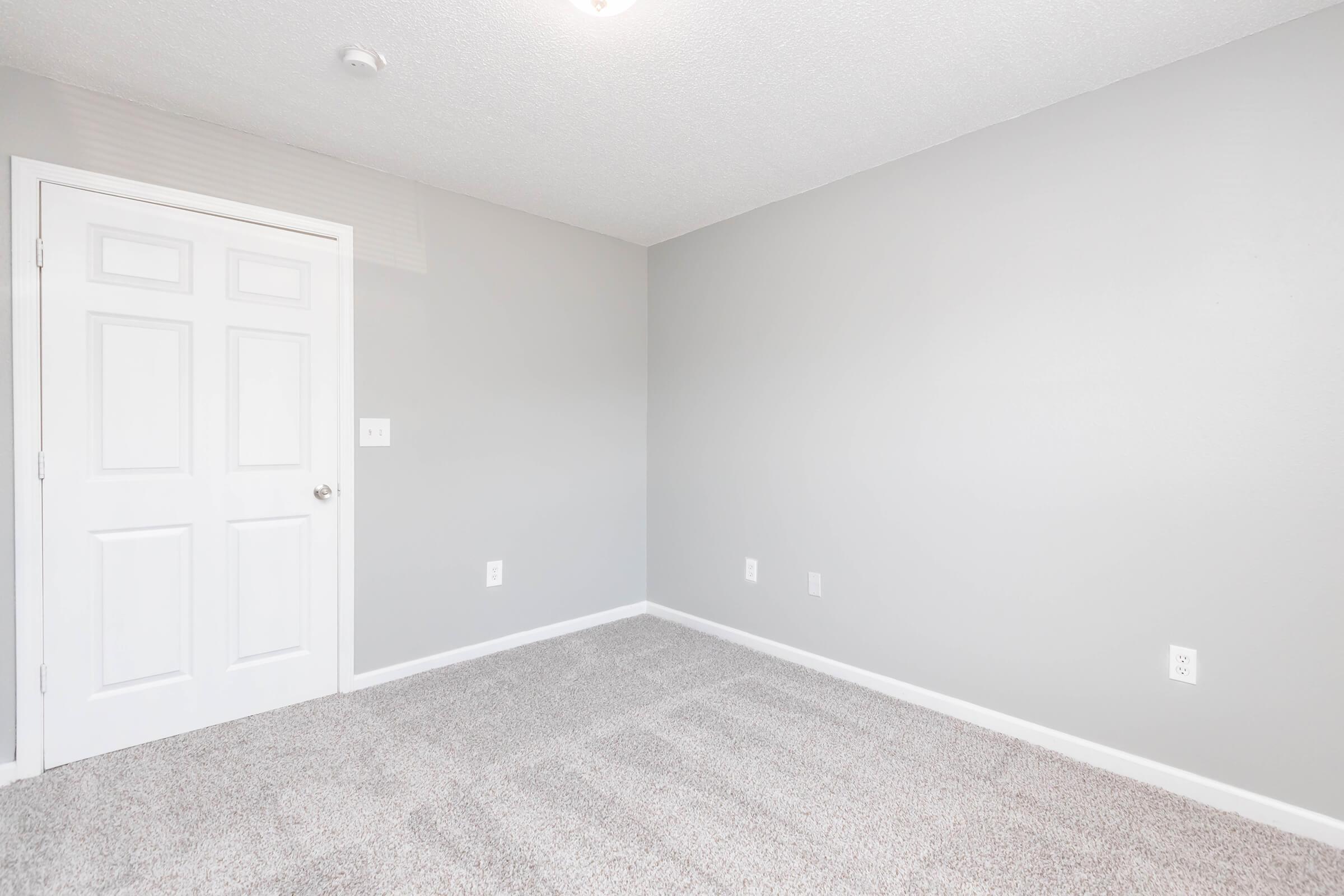 A small, empty room with light gray walls and a carpeted floor. There is a white door on the left and a ceiling light. The room has no furniture, creating an open space with clean lines and a minimalist feel.