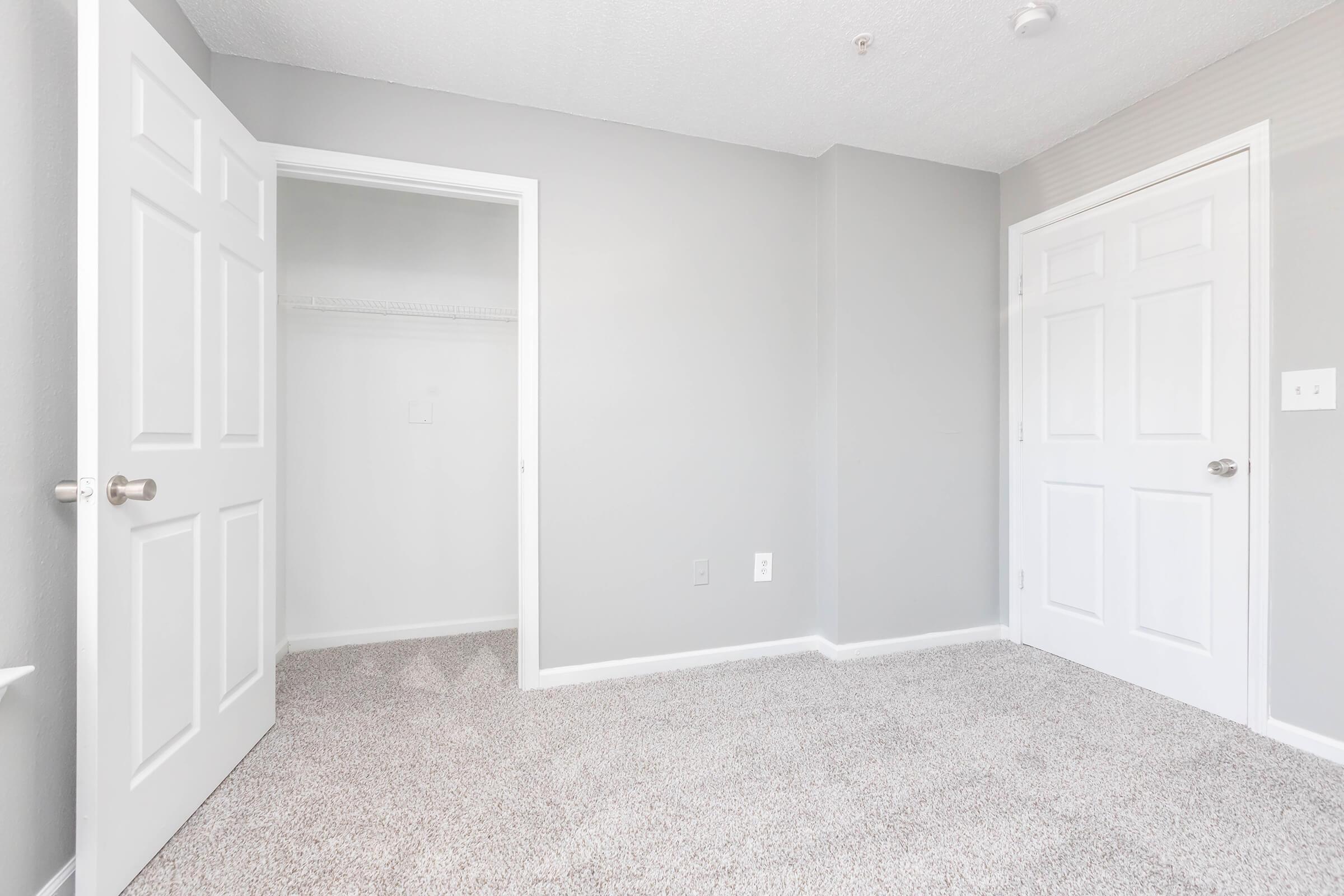 A clean, empty bedroom featuring light gray walls, plush carpet, and two white doors. One door leads to a closet, while the other opens to an adjoining room. The space is well-lit, creating a feeling of openness and tranquility.