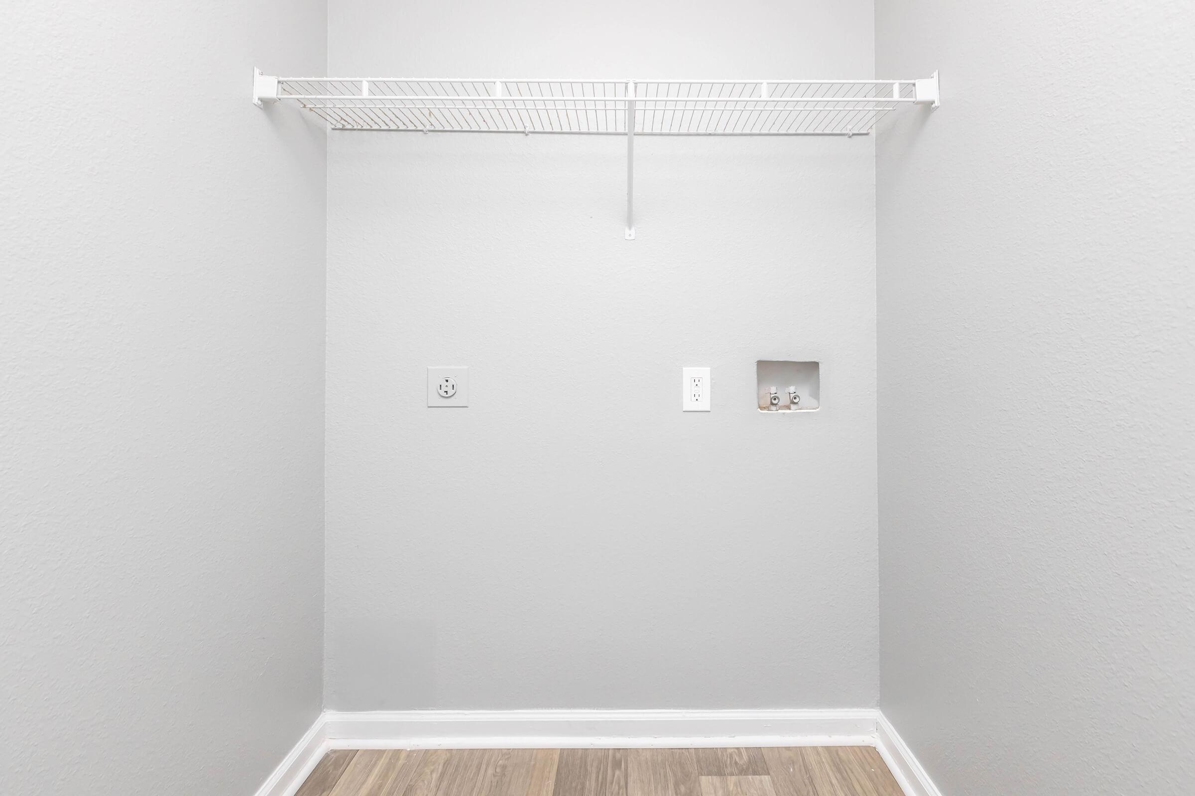 An empty, light gray closet with a wire shelf mounted on the back wall. The flooring is wood-look laminate. There are two electrical outlets and a small recessed alcove on the wall, with no visible items stored in the space.
