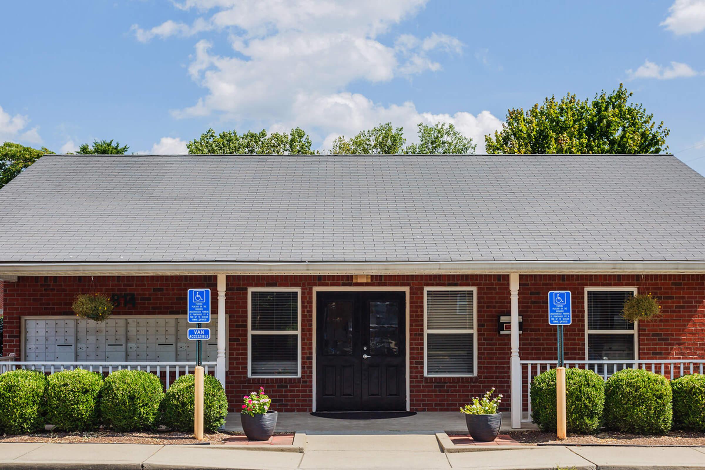 A brick building with a gray roof and a black front door. The entrance features white railings and is flanked by small bushes. Two accessible parking signs are visible in front, along with mailbox units beside the door. The sky is clear with a few clouds.