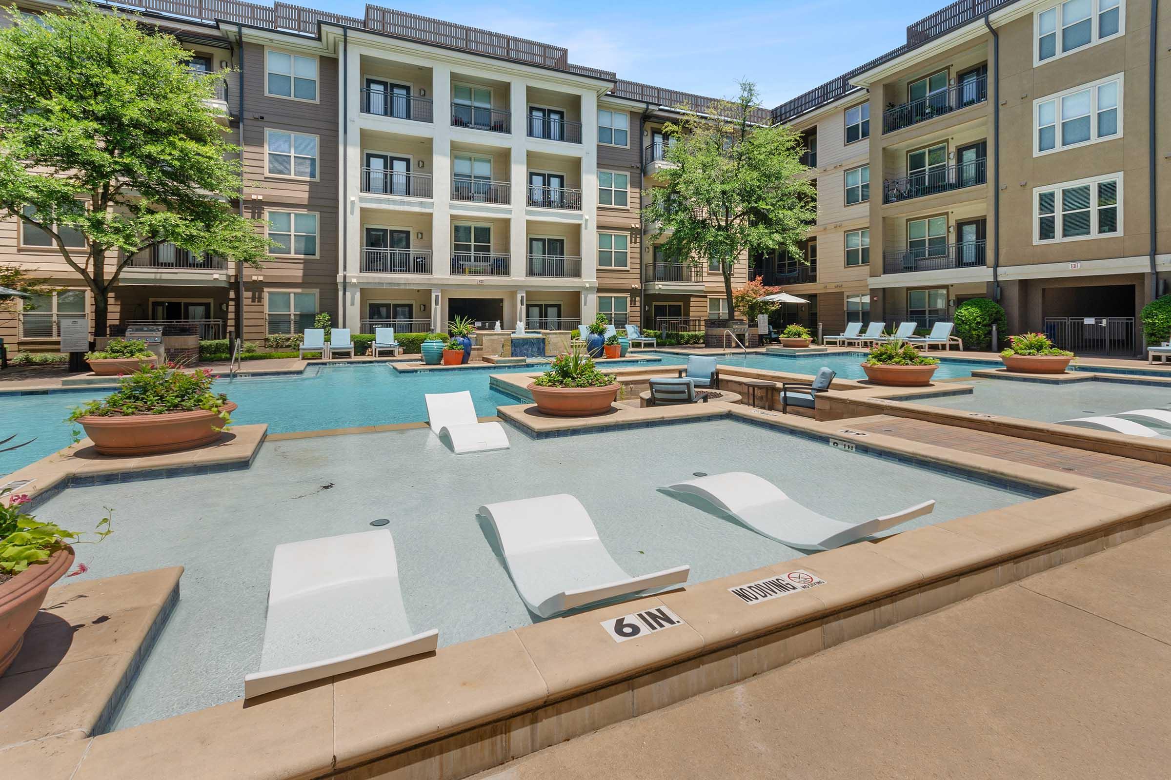 A modern apartment complex featuring a large outdoor pool with lounge chairs arranged around it. Surrounded by well-maintained landscaping, potted plants, and multiple seating areas, the pool area offers a relaxing atmosphere for residents.