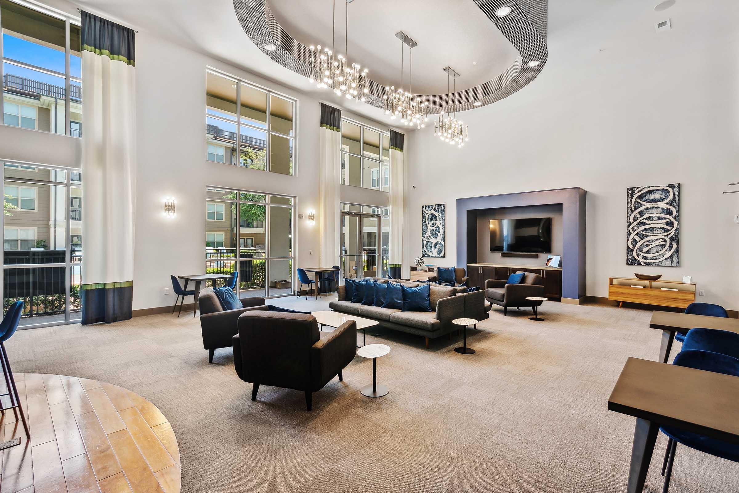 A spacious and modern lounge area featuring a stylish seating arrangement with blue and black couches, elegant light fixtures, large windows allowing natural light, and contemporary artwork on the walls.