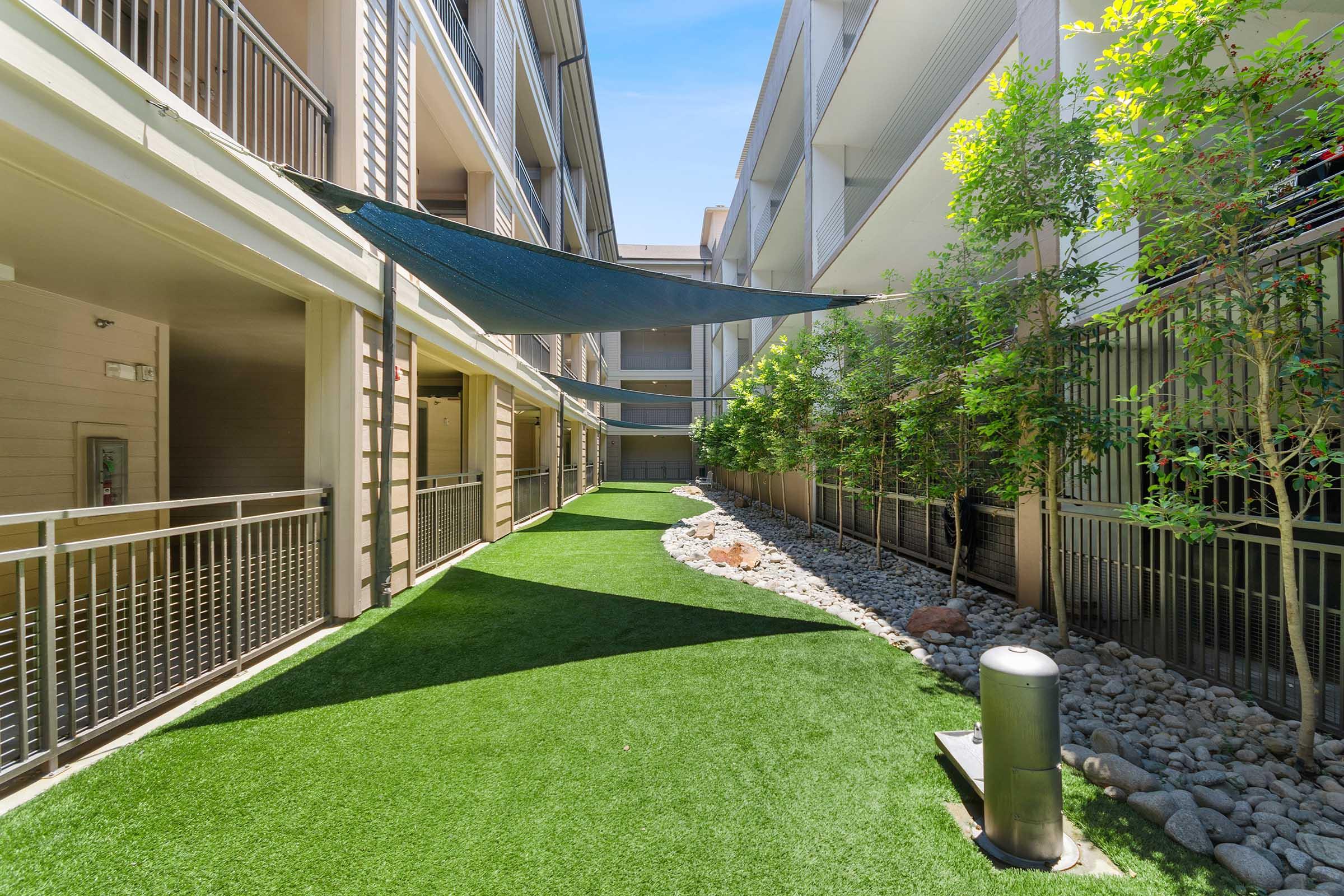 Lush green artificial turf pathway between modern apartment buildings, lined with small rocks and plants.