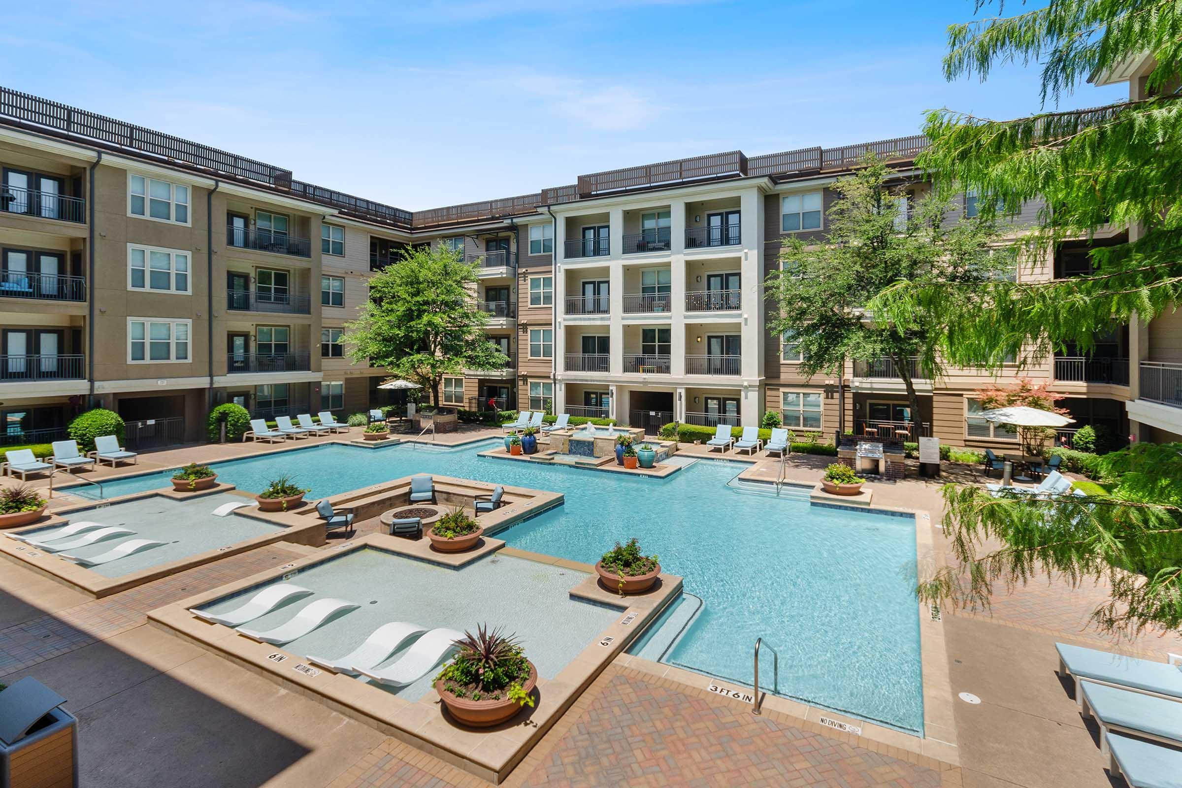 A spacious and modern apartment complex featuring a central swimming pool surrounded by lounge chairs and landscaped planters.