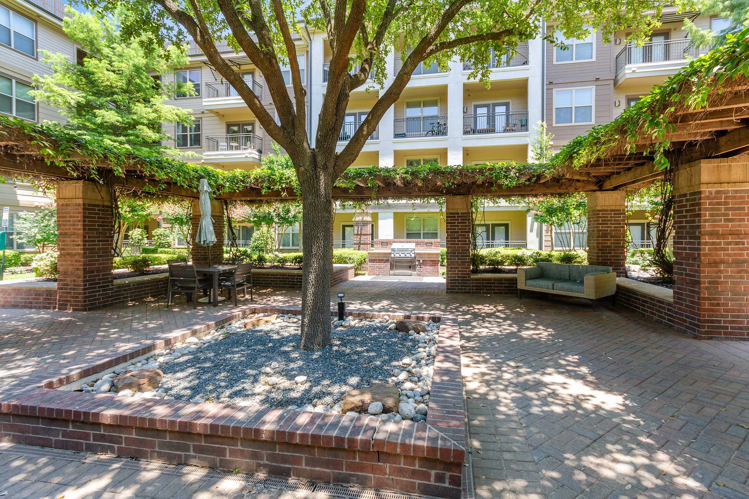 A landscaped outdoor space featuring a central tree surrounded by a brick-bordered garden, with a patio area that includes seating and a grill.