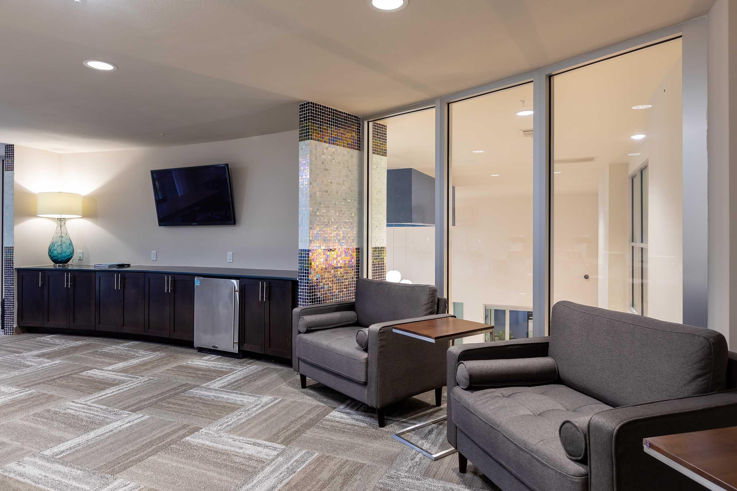 A modern lounge area featuring two gray armchairs with side tables, a flat-screen TV mounted on the wall, and a decorative light fixture.