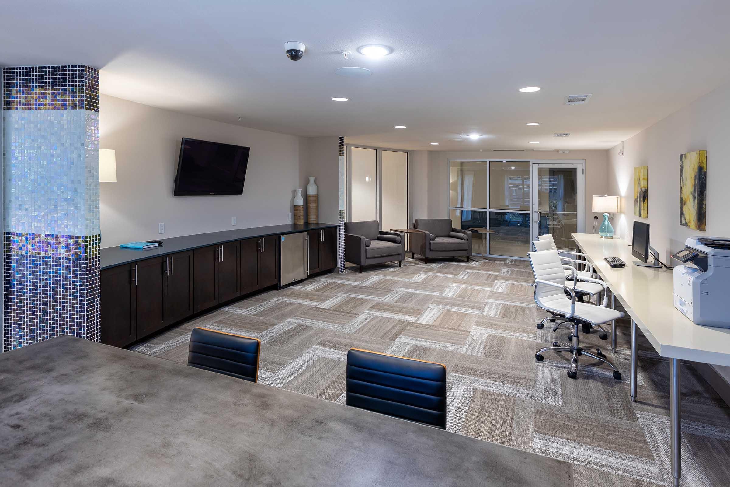 Modern interior of a commercial space featuring a reception area with a large flat-screen TV, two armchairs, a desk with multiple office chairs, and computers.
