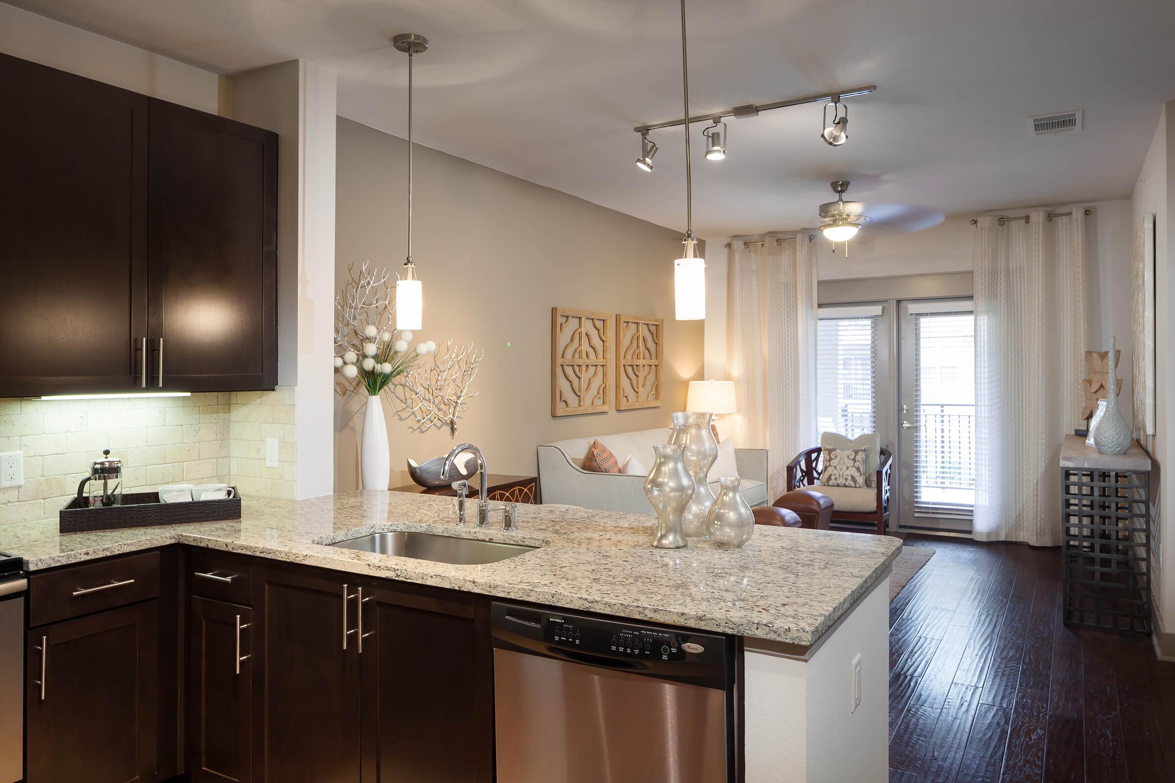A modern kitchen with dark cabinets and granite countertops, featuring a sink and stainless steel appliances. Pendant lights hang above the counter.