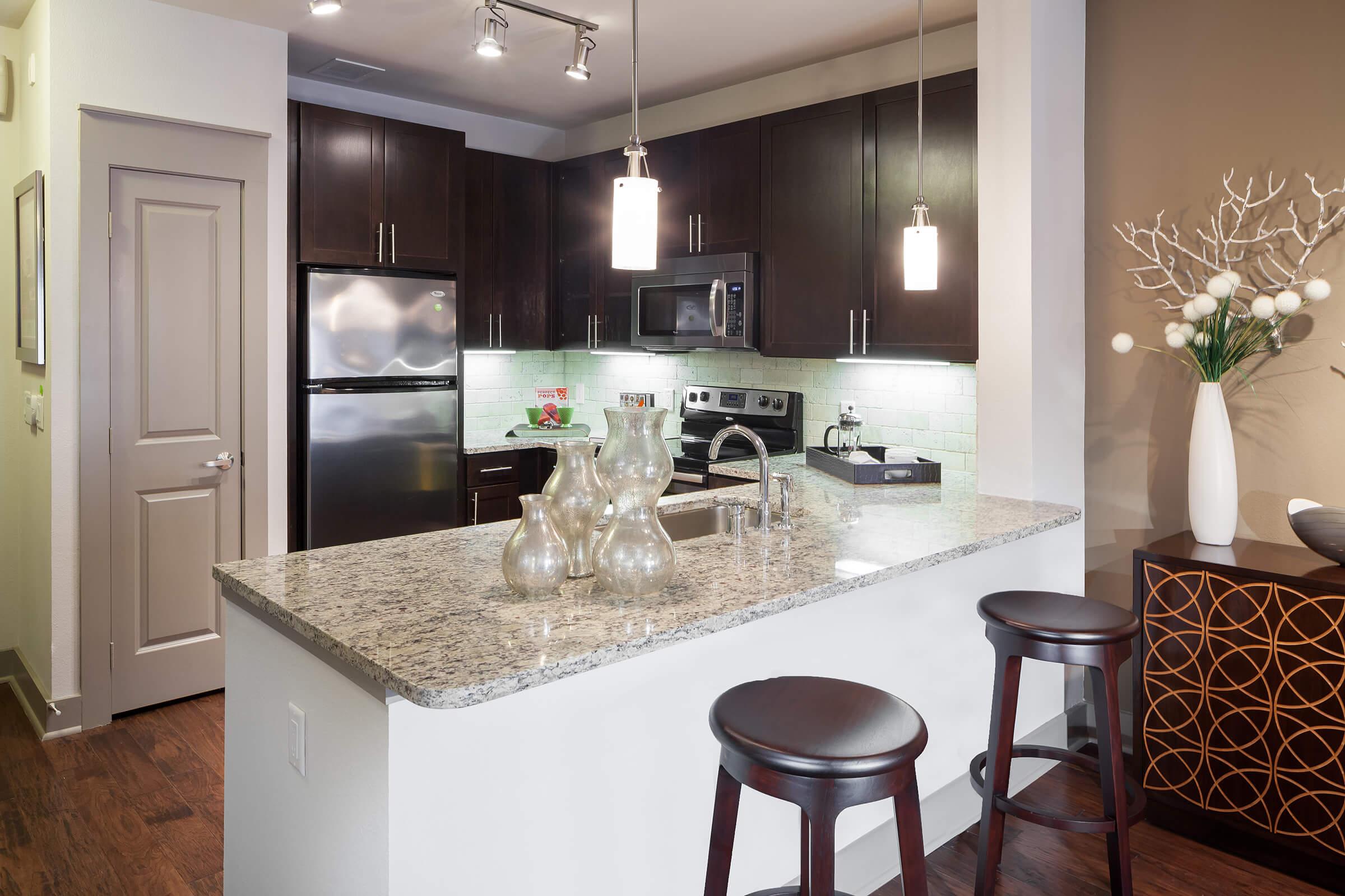Modern kitchen featuring dark wood cabinets, a granite countertop with decorative vases, stainless steel appliances, and a small dining area with two wooden stools.