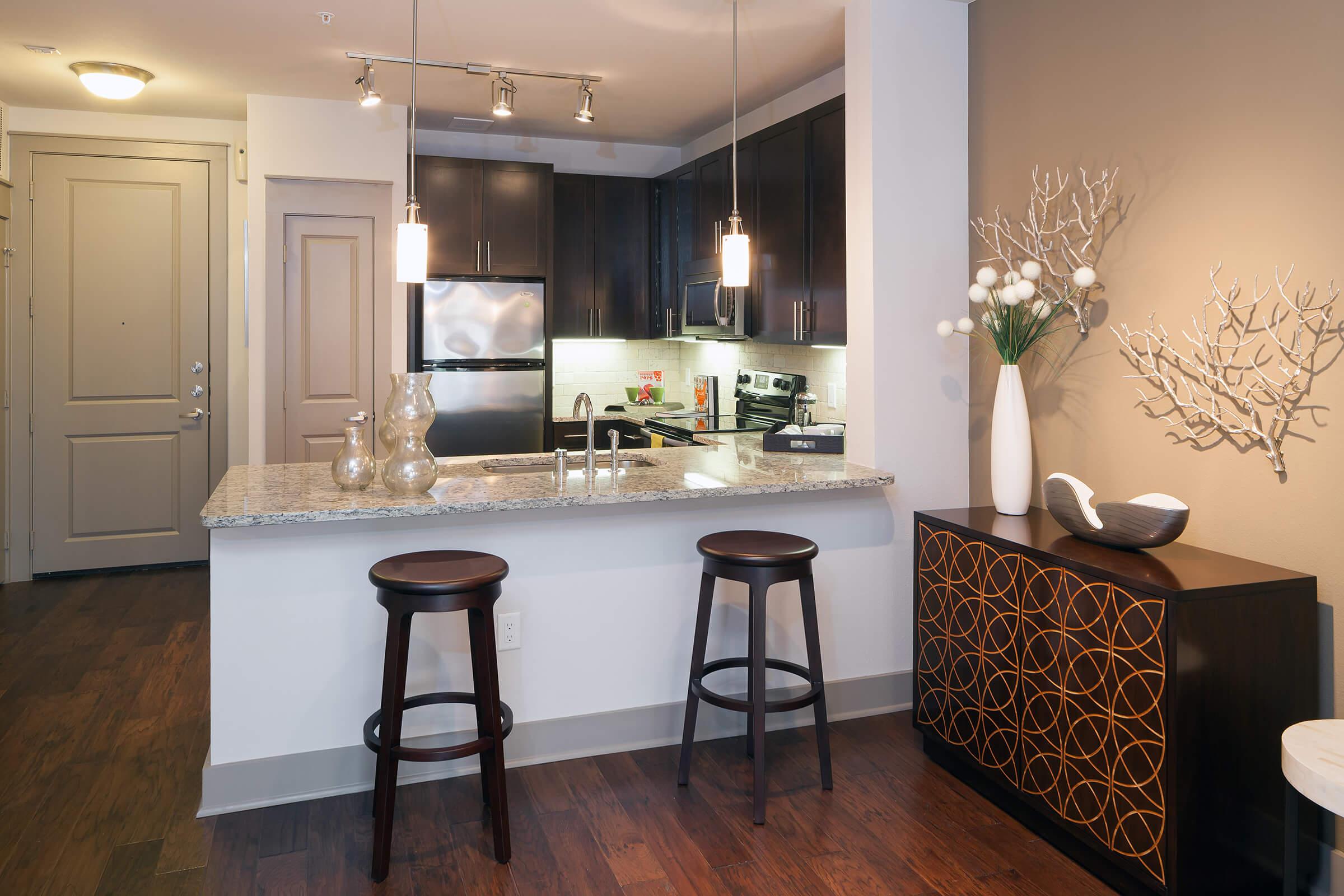 Modern kitchen and dining area featuring a granite countertop with two bar stools, stainless steel appliances, and stylish pendant lighting.