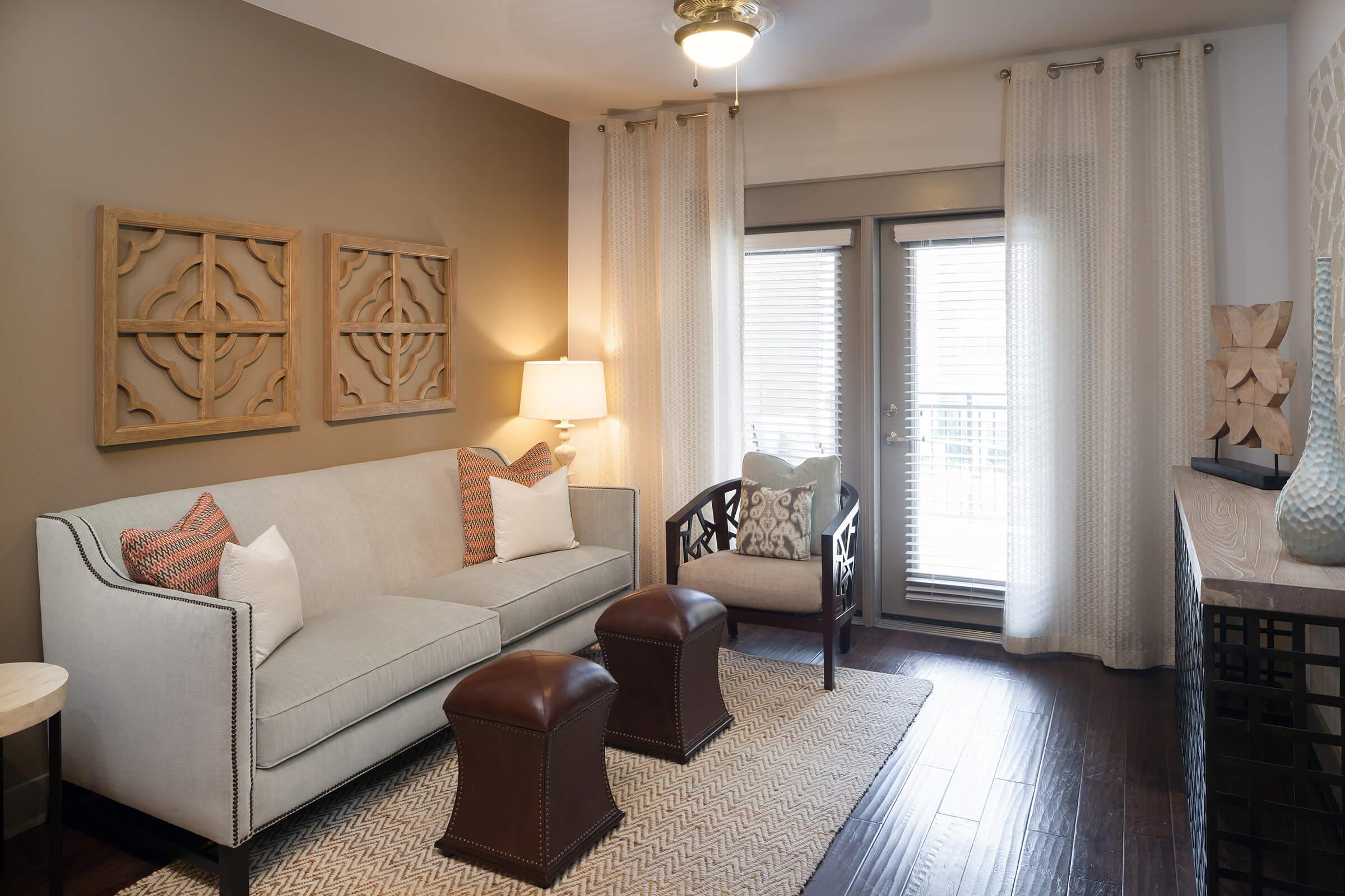Cozy living room featuring a light gray sofa with decorative pillows, a wooden chair with a patterned cushion, and two brown ottomans.
