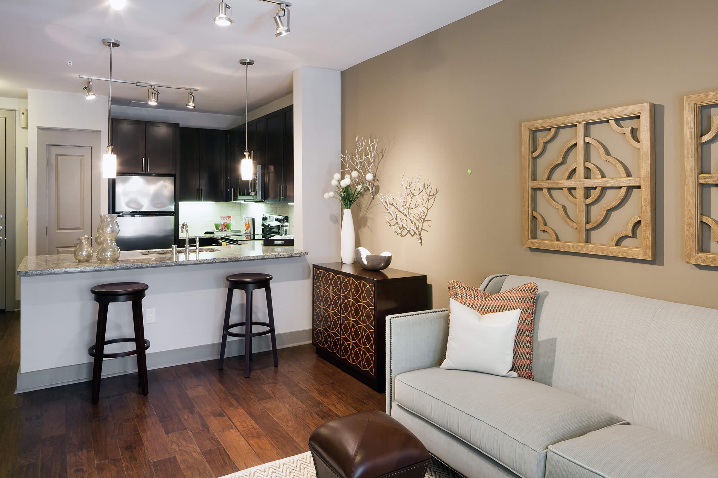 Modern living room and kitchen in an apartment. Features a sofa with decorative pillows, a stylish coffee table, and a kitchen with dark cabinetry and granite countertops.