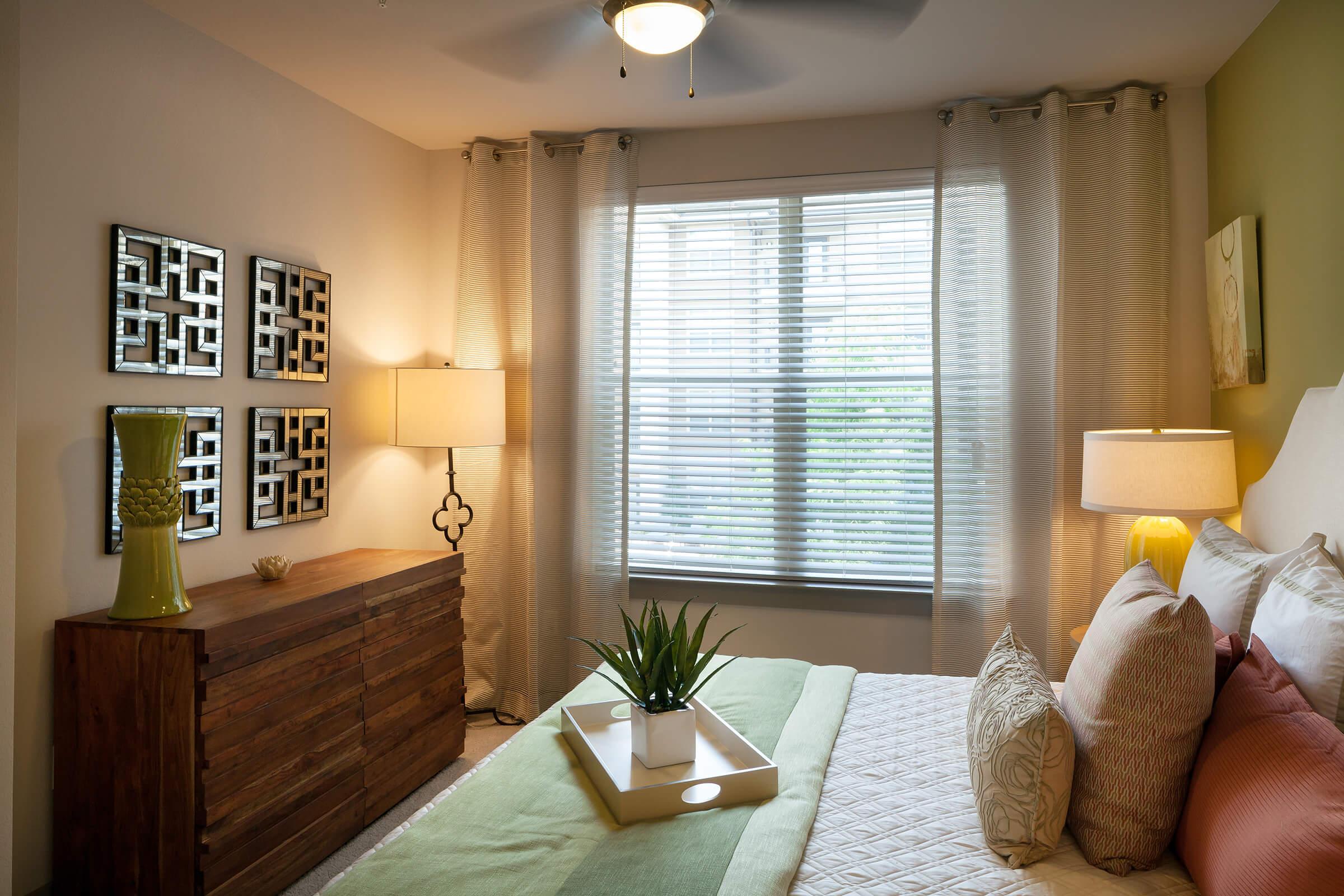 A cozy bedroom featuring a queen-sized bed with decorative pillows, a wooden dresser, two table lamps, and a window with sheer curtains.