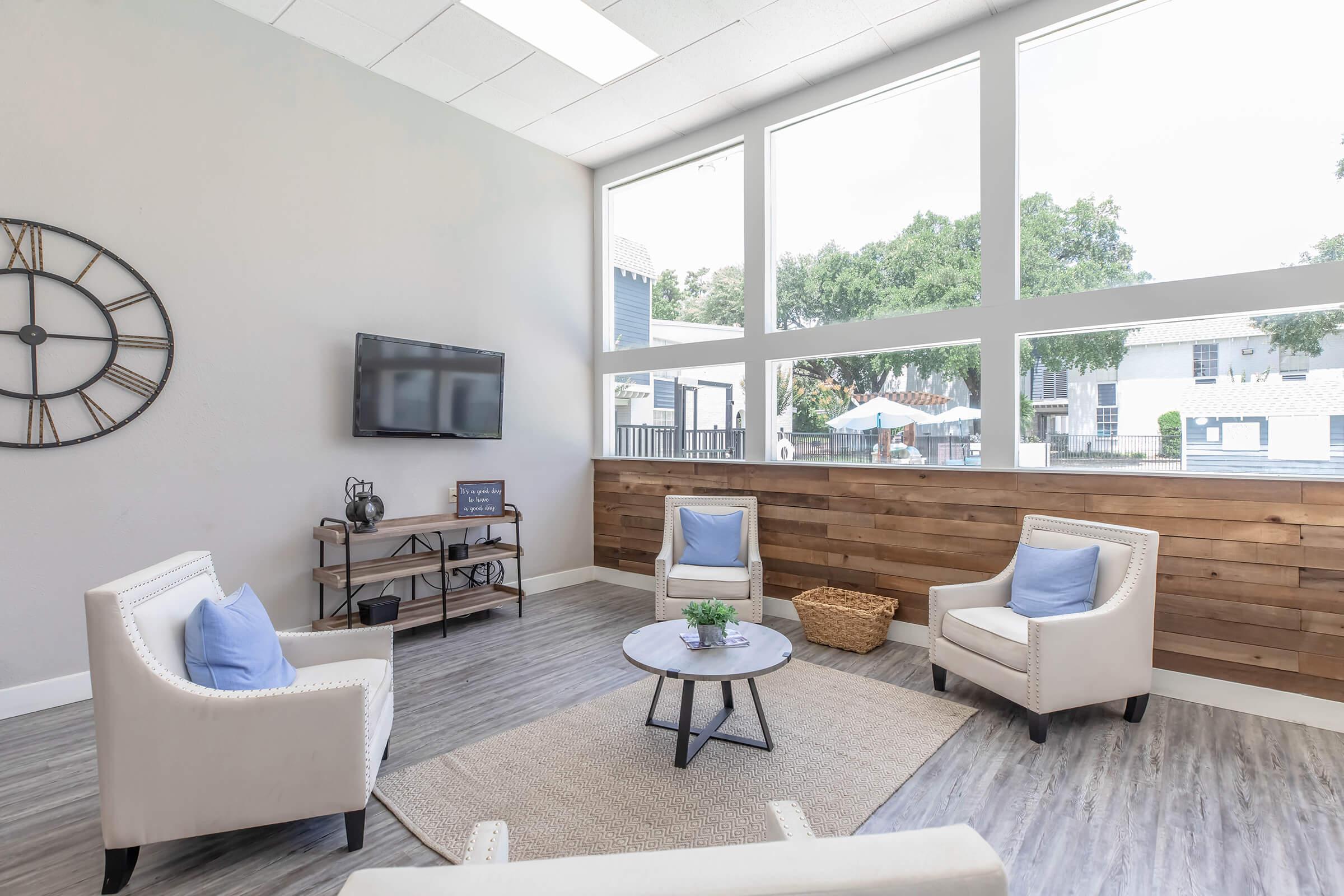 A modern lounge area featuring two white armchairs with blue cushions, a round coffee table, and a decorative plant. The room has wooden accents and large windows allowing natural light in. A wall clock and a TV are visible, creating a welcoming atmosphere.