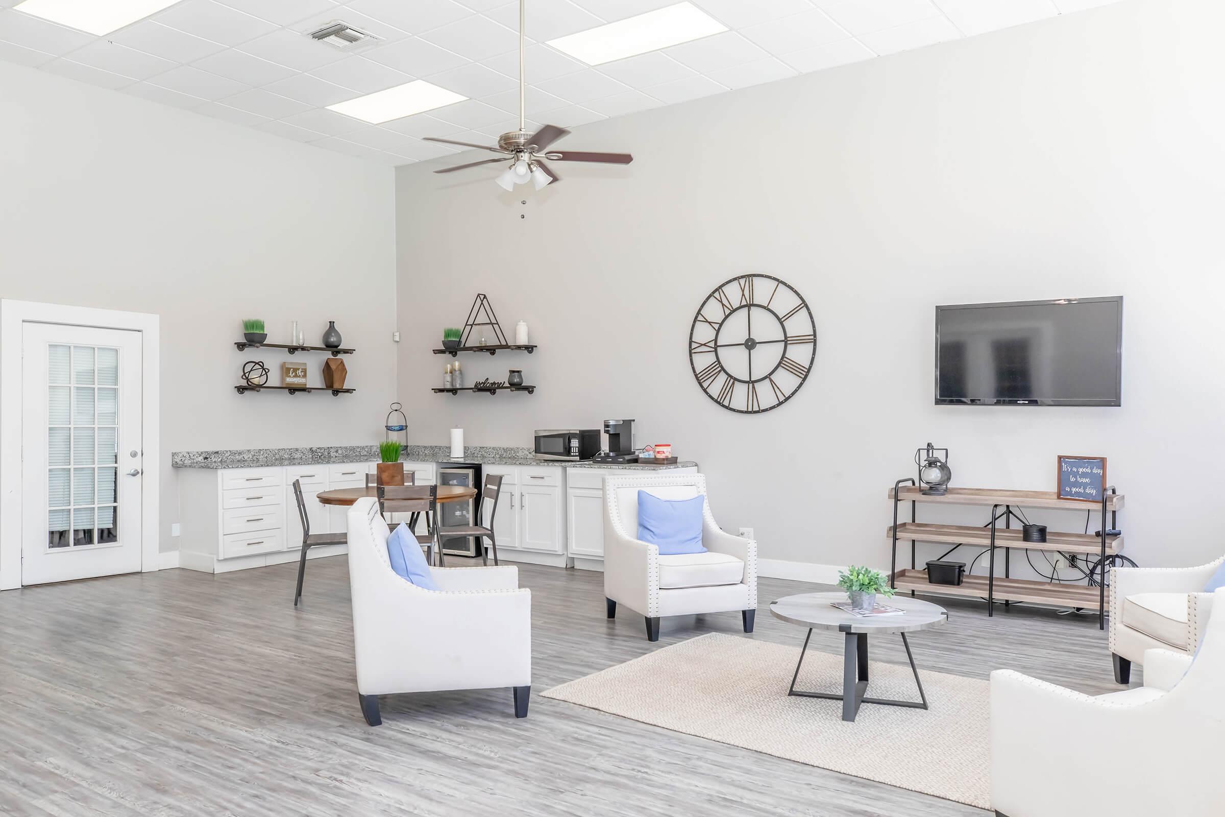 A bright and airy lounge area featuring two white upholstered chairs with blue cushions, a round table, and a television mounted on the wall. A large wall clock and shelving with decorative items add to the decor. There’s a kitchenette area with a countertop, microwave, and additional seating.