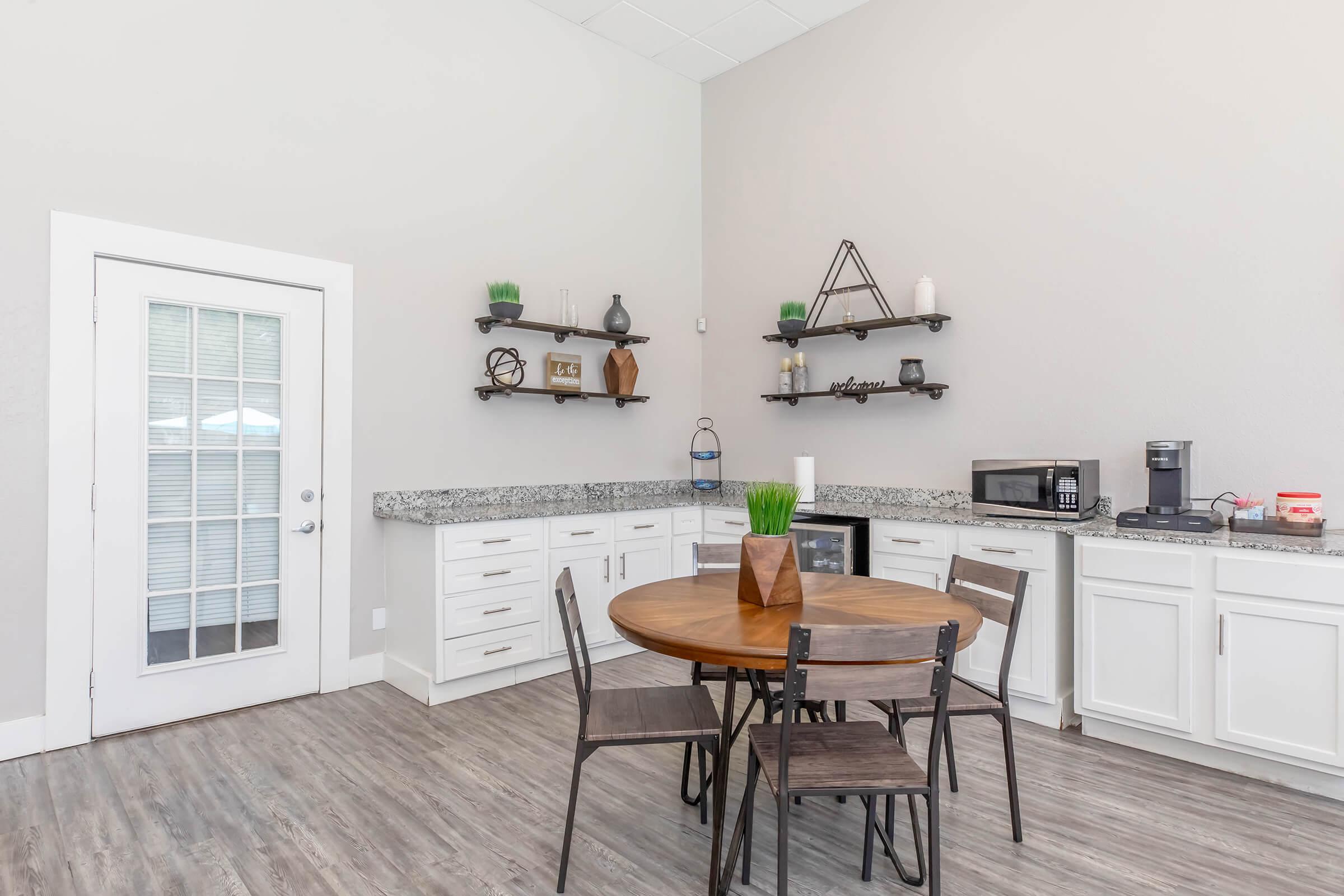A modern kitchen with a round wooden dining table and four wooden chairs. There are white cabinets and a grey granite countertop. Shelves on the wall display decorative items. A microwave and coffee maker are visible, along with a glass door leading to an exterior area. The flooring is light-colored wood.