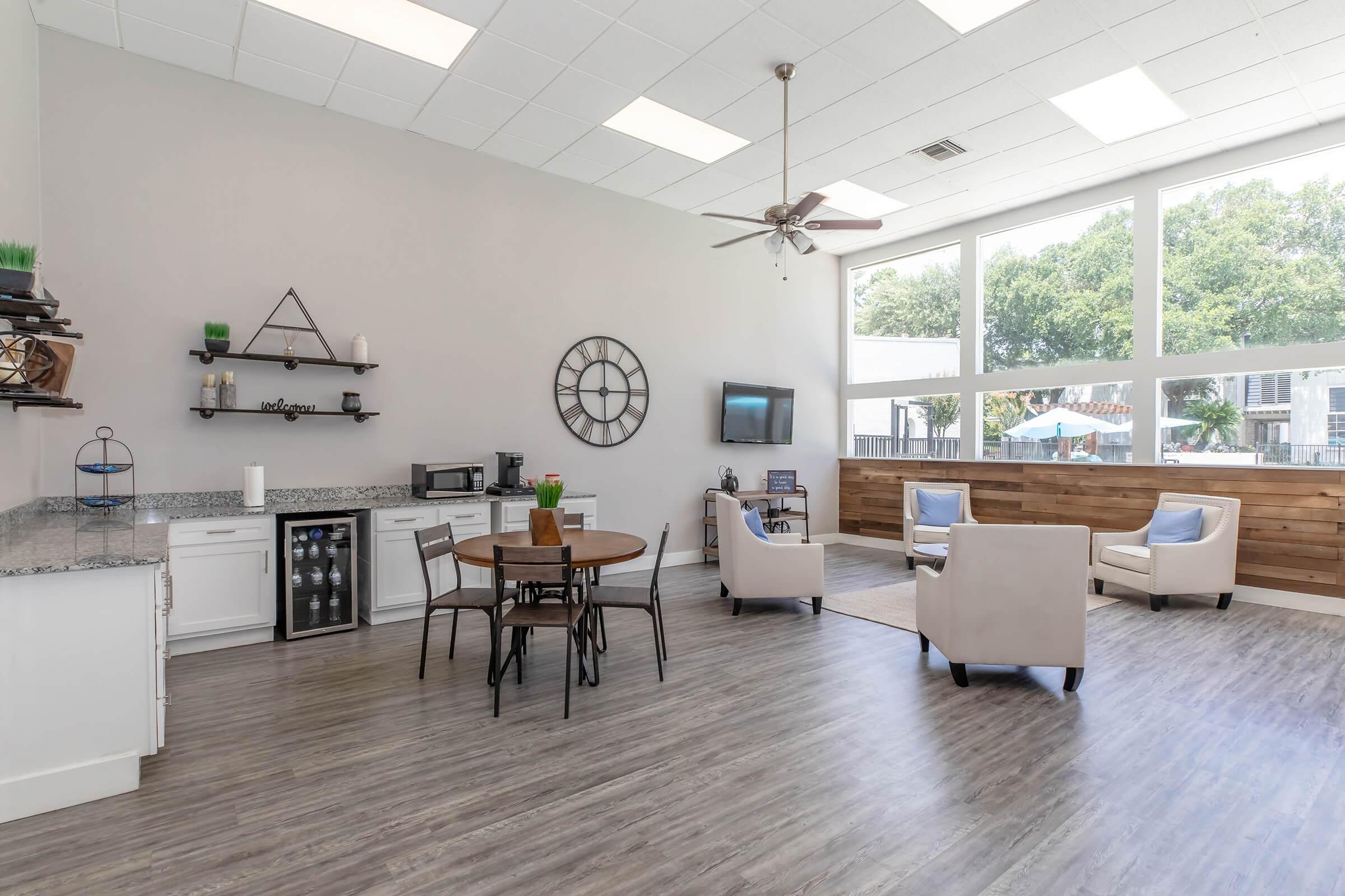 Bright and spacious common area featuring a round dining table with chairs, a kitchenette with a microwave and refrigerator, a large wall clock, and comfortable seating arrangements. Large windows allow plenty of natural light, with a view of greenery outside. The decor is modern and inviting.