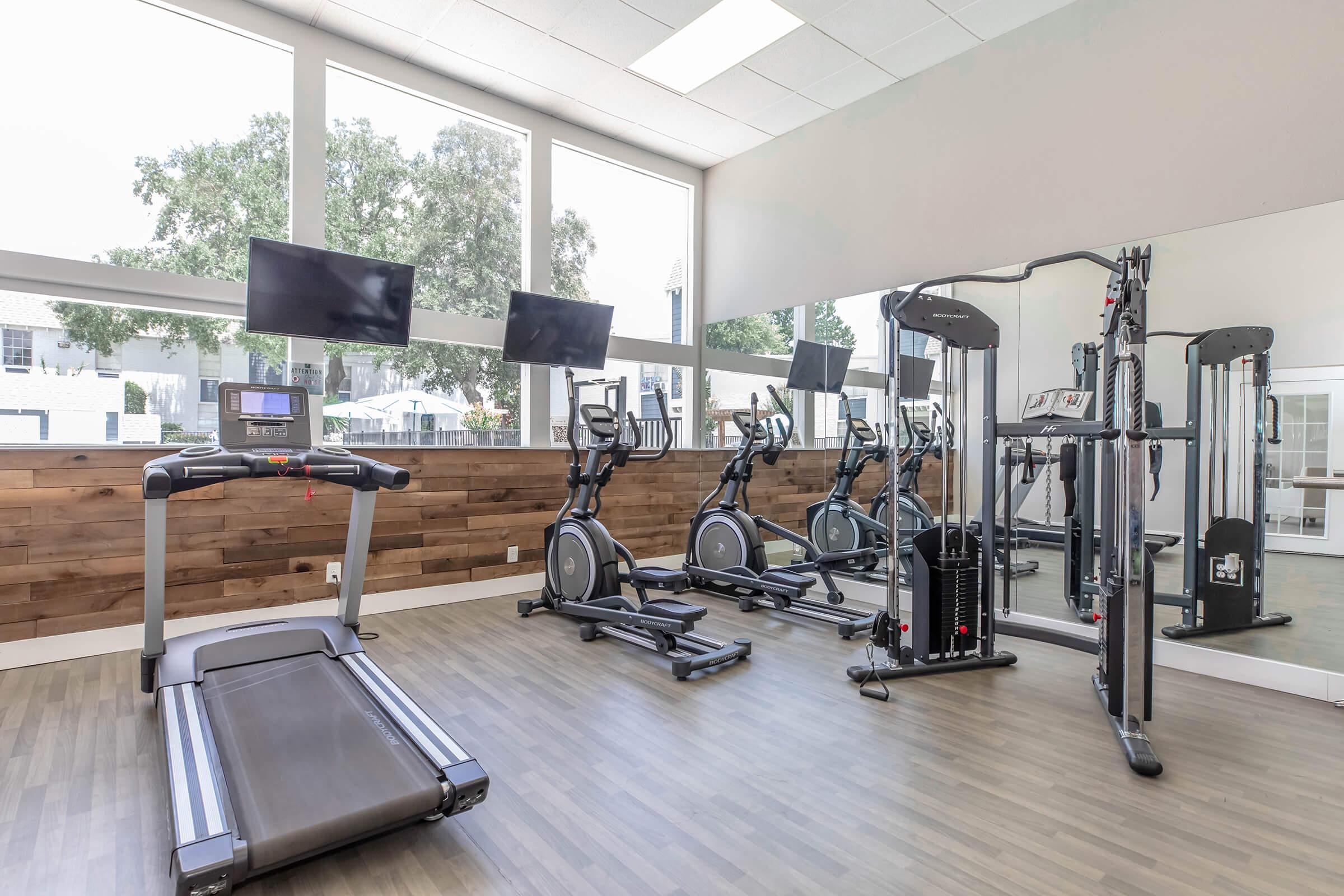 A modern fitness gym with a treadmill, elliptical machines, and a multi-gym set. The room features large windows allowing natural light, wooden accents on the wall, and two flat-screen TVs mounted for entertainment. The flooring is a sleek, light-colored wood.