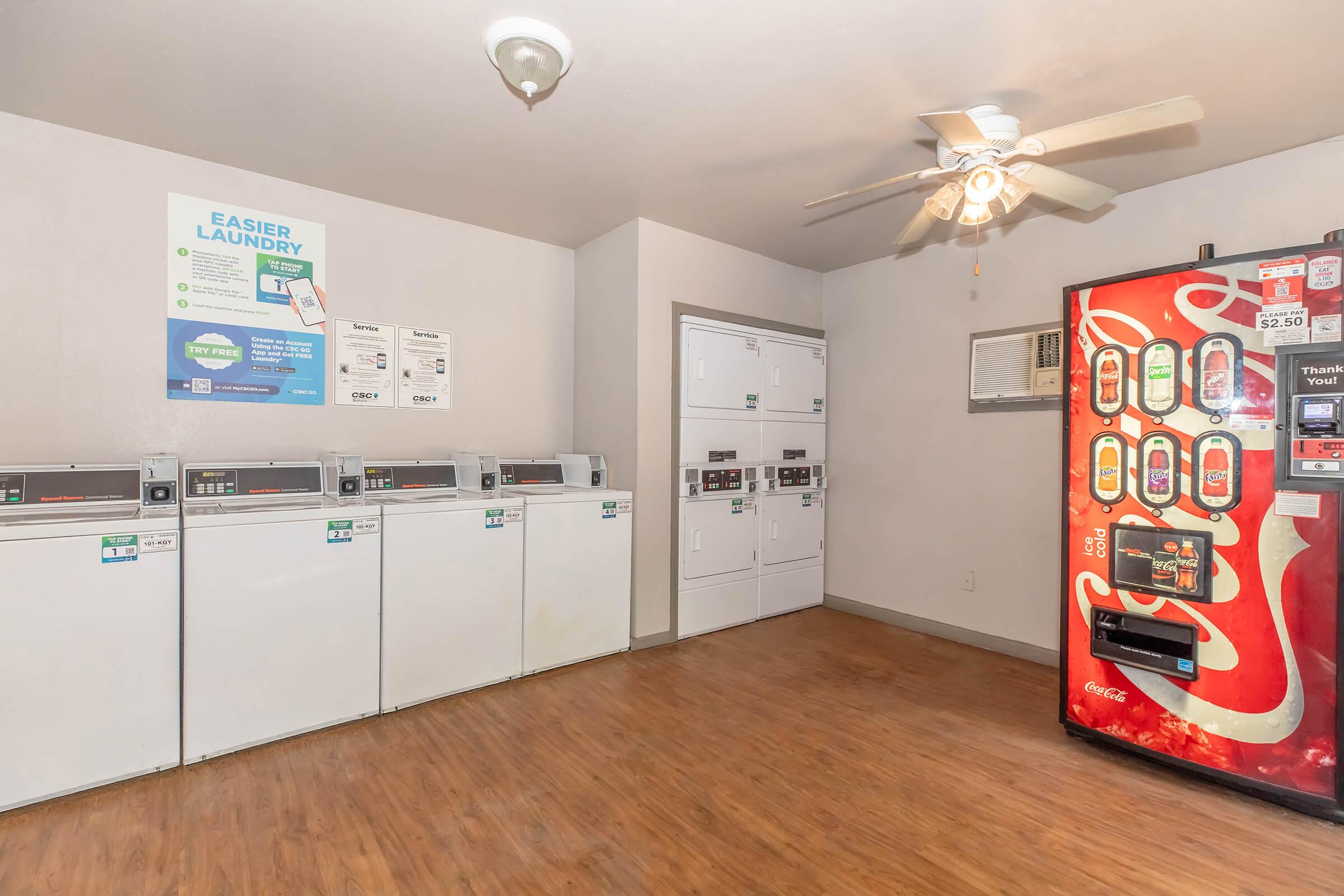 A clean laundry room featuring rows of white washing machines and dryers, an air conditioning unit, and a Coca-Cola vending machine. The walls are light-colored, and a ceiling fan is visible. Laundry instructions and service information are posted on the wall.