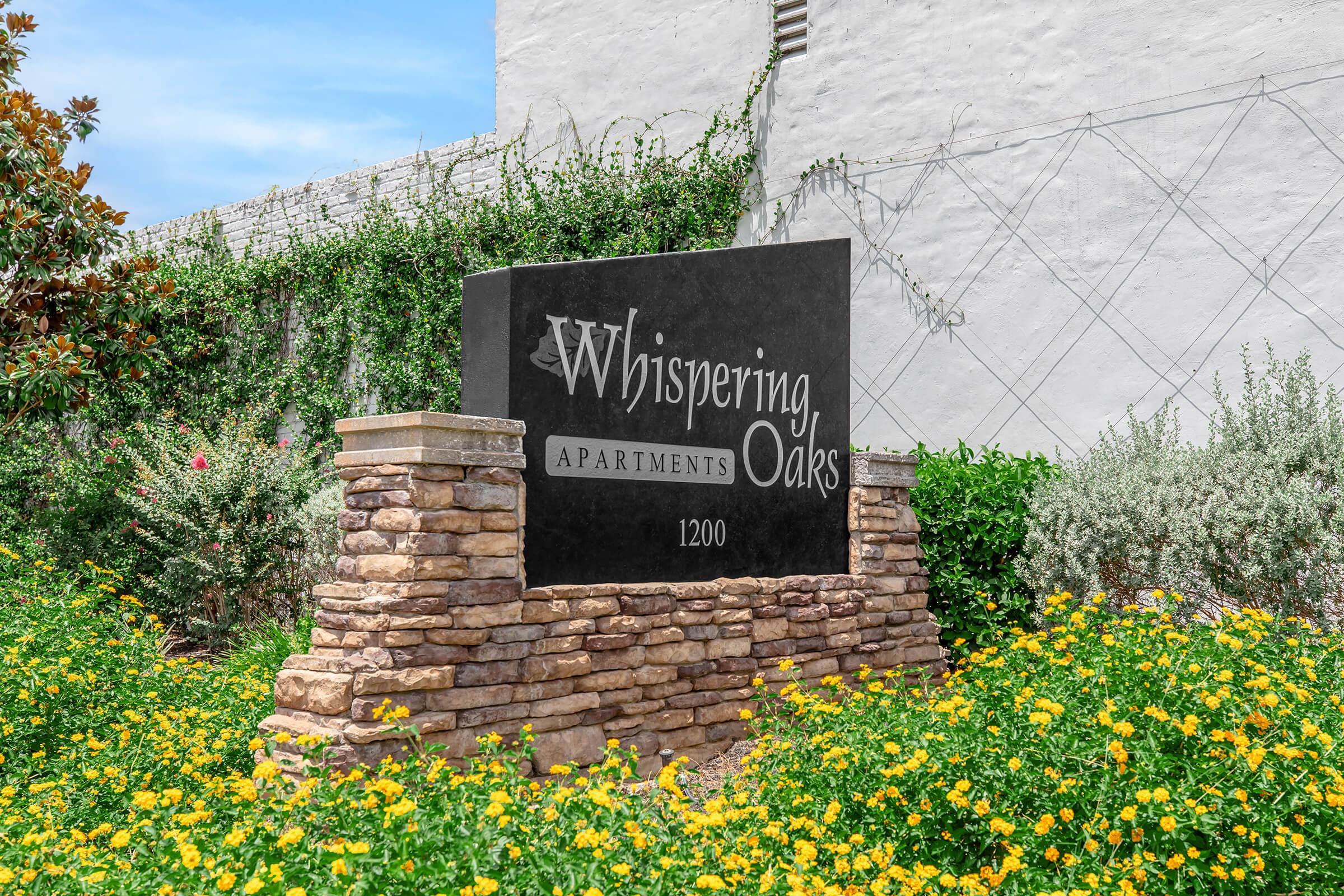 Sign for Whispering Oaks Apartments, featuring elegant lettering on a dark stone base. Surrounded by vibrant yellow flowers and lush greenery, enhancing the welcoming atmosphere. The location indicated is 1200, with a background of a white building and blue sky.
