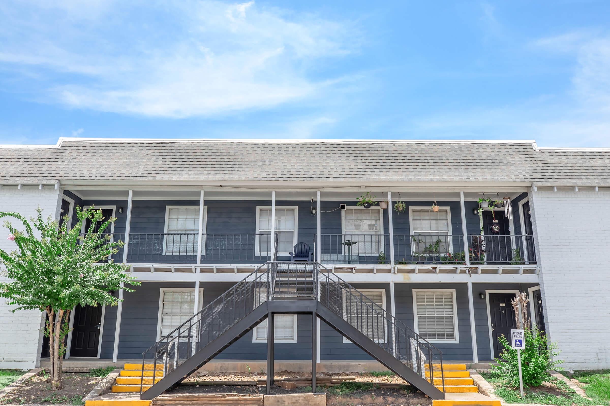 Two-story apartment building with a gray exterior and white trim. The front features a set of stairs leading to the second floor, with a balcony adorned by potted plants. The sky is bright blue with a few clouds, and there are trees and a small garden area in the foreground.