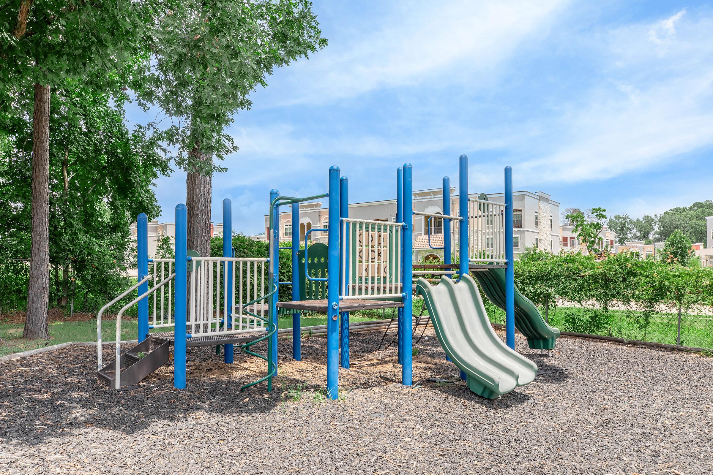 A colorful playground featuring climbing structures, slides, and a spiral slide, surrounded by grassy areas and trees. The sunny sky and nearby buildings provide a cheerful backdrop, inviting children to play and explore.