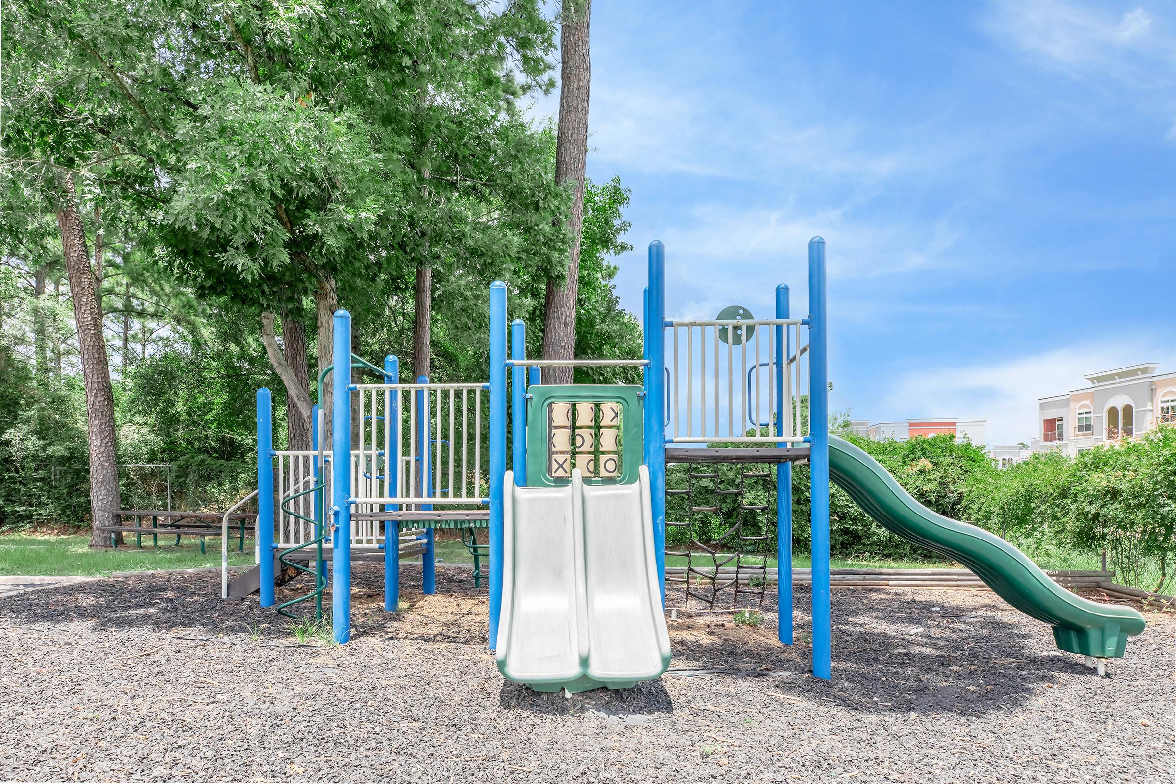 A playground featuring two slides, climbing structures, and safety railings, situated on a bed of wood chips. Surrounding the playground are lush green trees and a clear blue sky, creating a bright and inviting outdoor space for children to play.