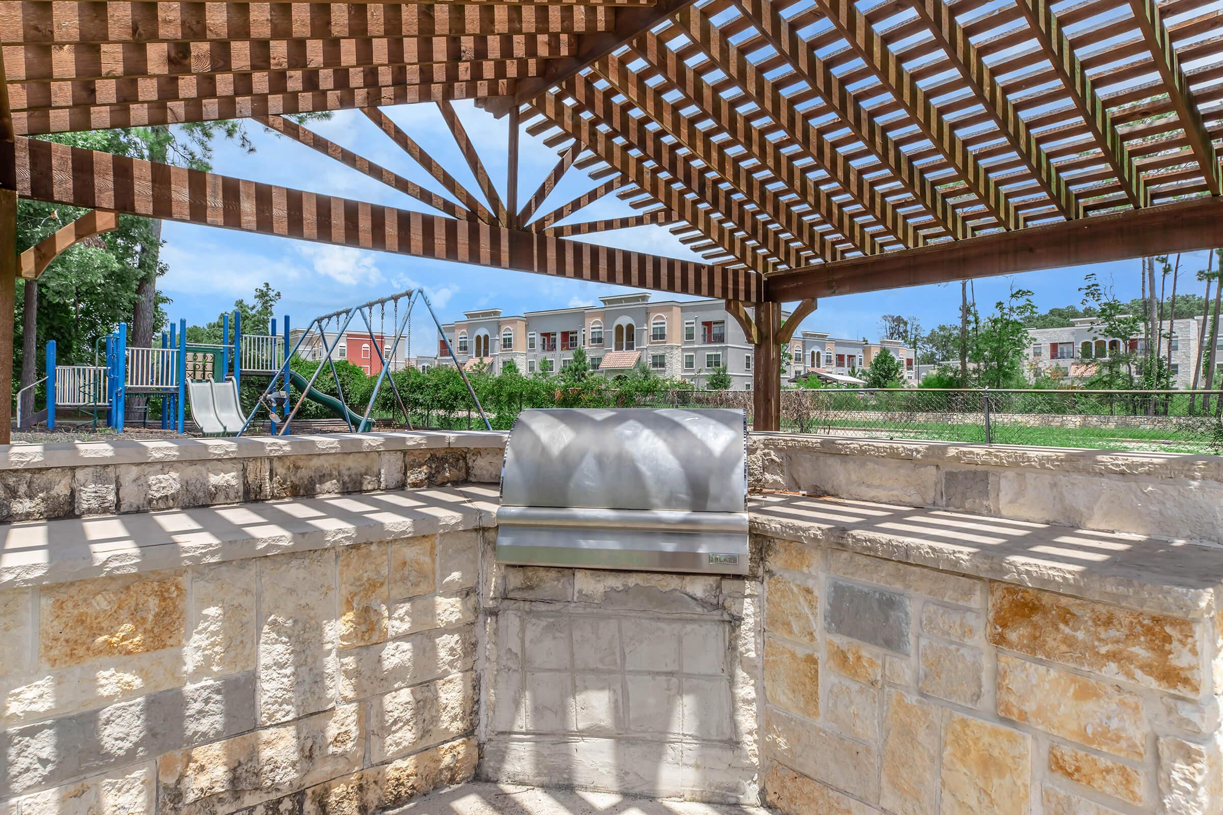 A covered outdoor space with wooden beams and a stone wall. In the background, there are swings and playground equipment, along with residential buildings and greenery. The area is bright and sunny, creating a welcoming atmosphere for outdoor activities and gatherings.